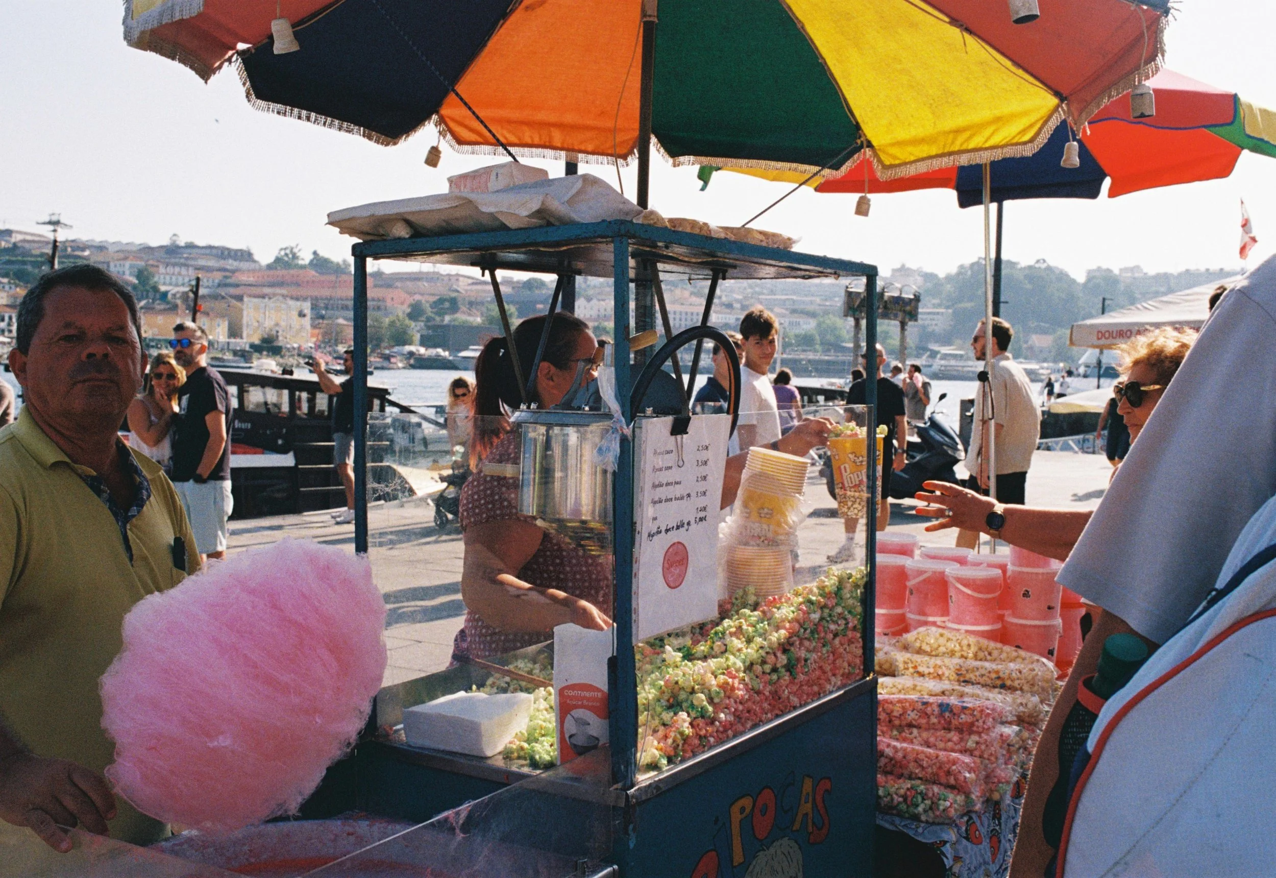 Un puesto de feria vendiendo algodón de azúcar y caramelos, con paraguas coloridos en un entorno junto al río.