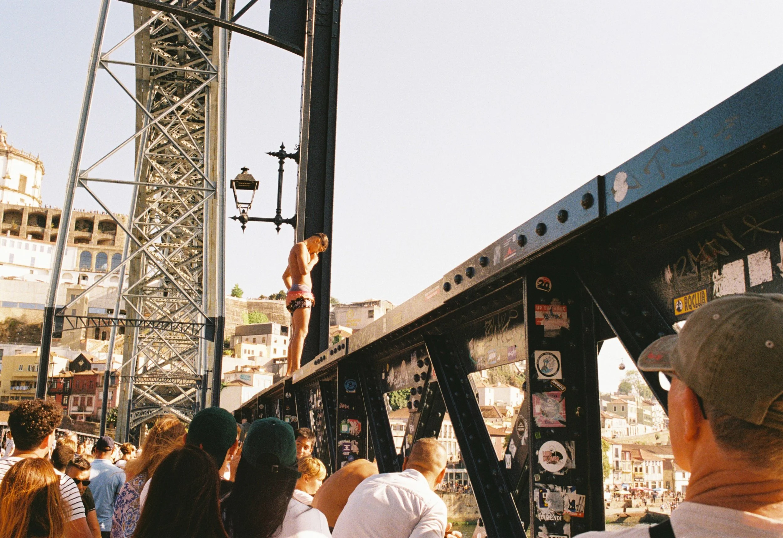 Joven en pantalones cortos y sin camiseta de pie sobre una estructura metálica, rodeada de espectadores, en una zona urbana con colinas y edificios en el fondo.