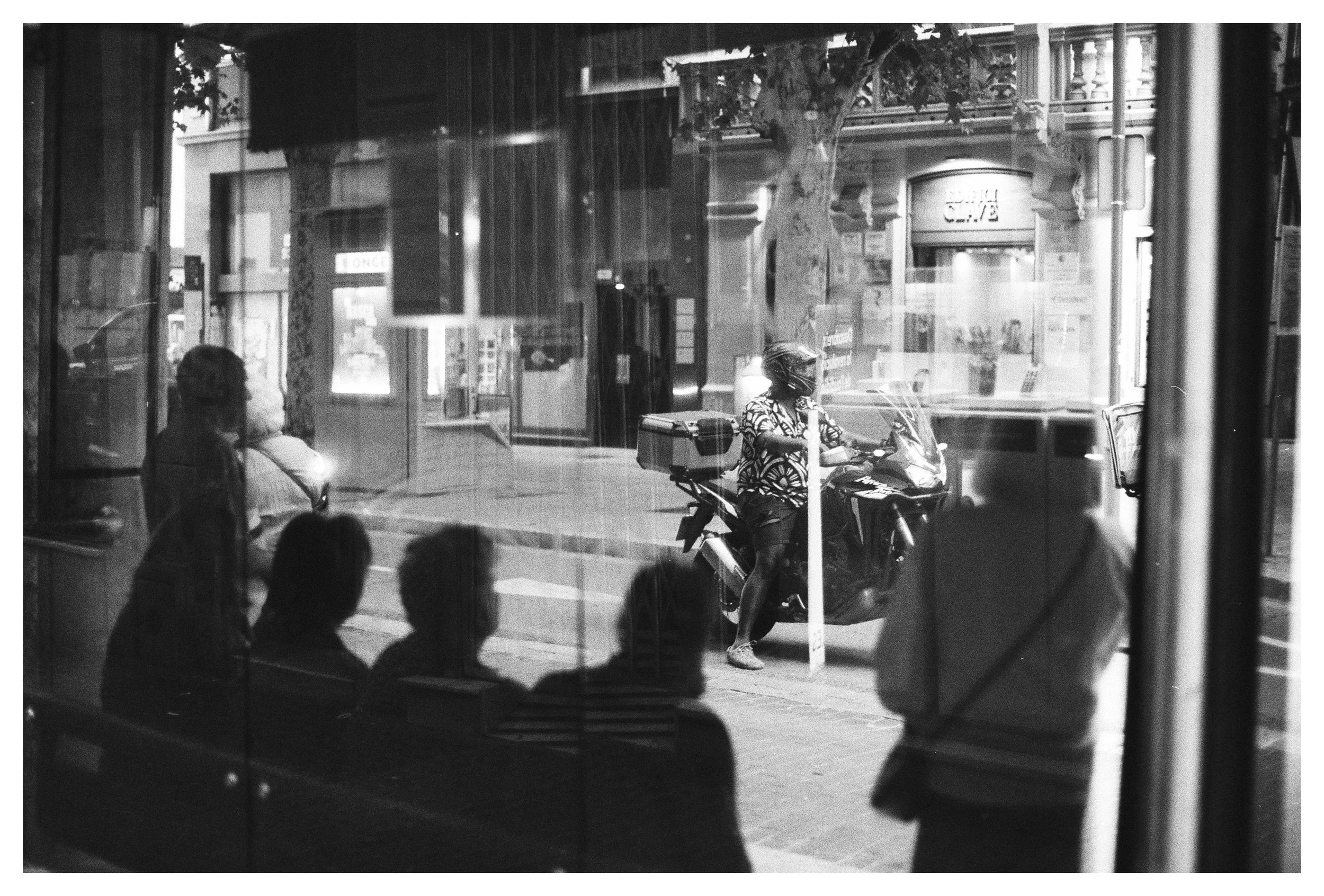 Imagen en blanco y negro vista desde el interior de un establecimiento, mostrando a varias personas sentadas y una persona con casco en una motocicleta del lado de la calle, y otra persona con casco sobre la motocicleta.