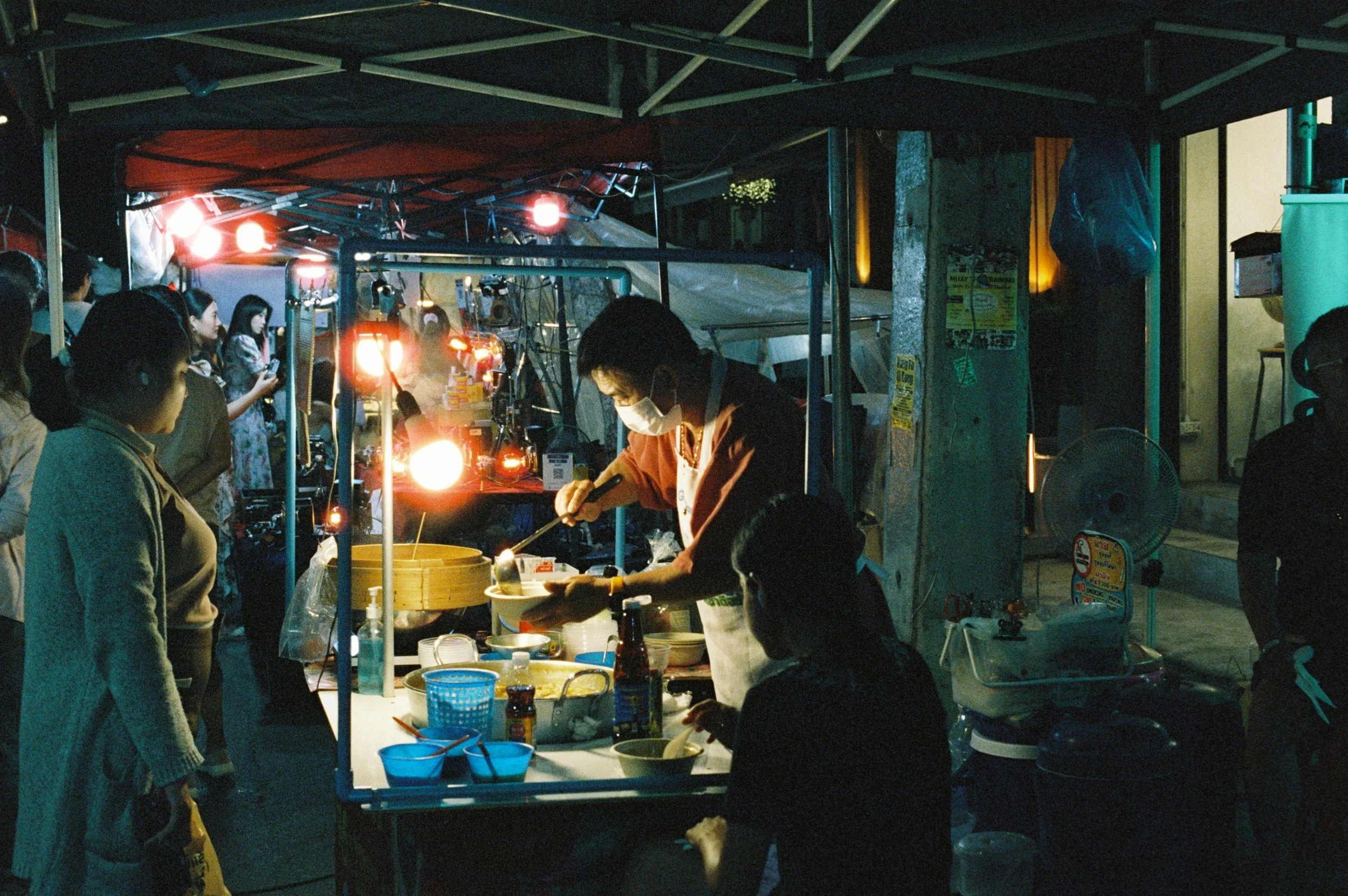 Vendedor preparando comida en un puesto callejero en la noche, con varias personas esperando ordenar. Iluminación artificial, ambiente nocturno.