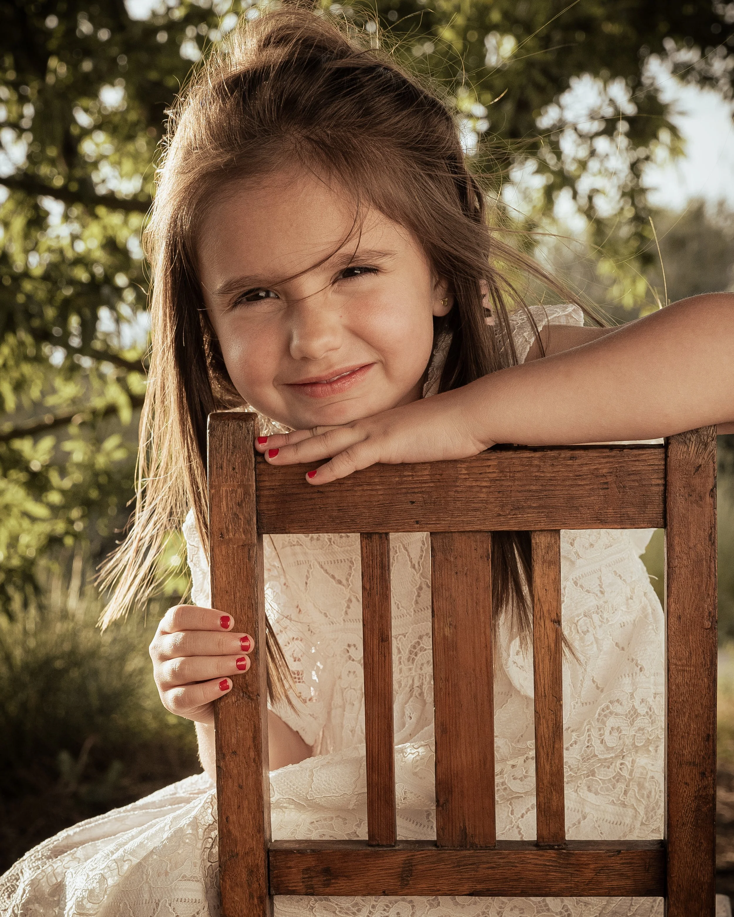 Niña sonriendo, descansando sobre una silla de madera en un entorno al aire libre con árboles y luz natural.