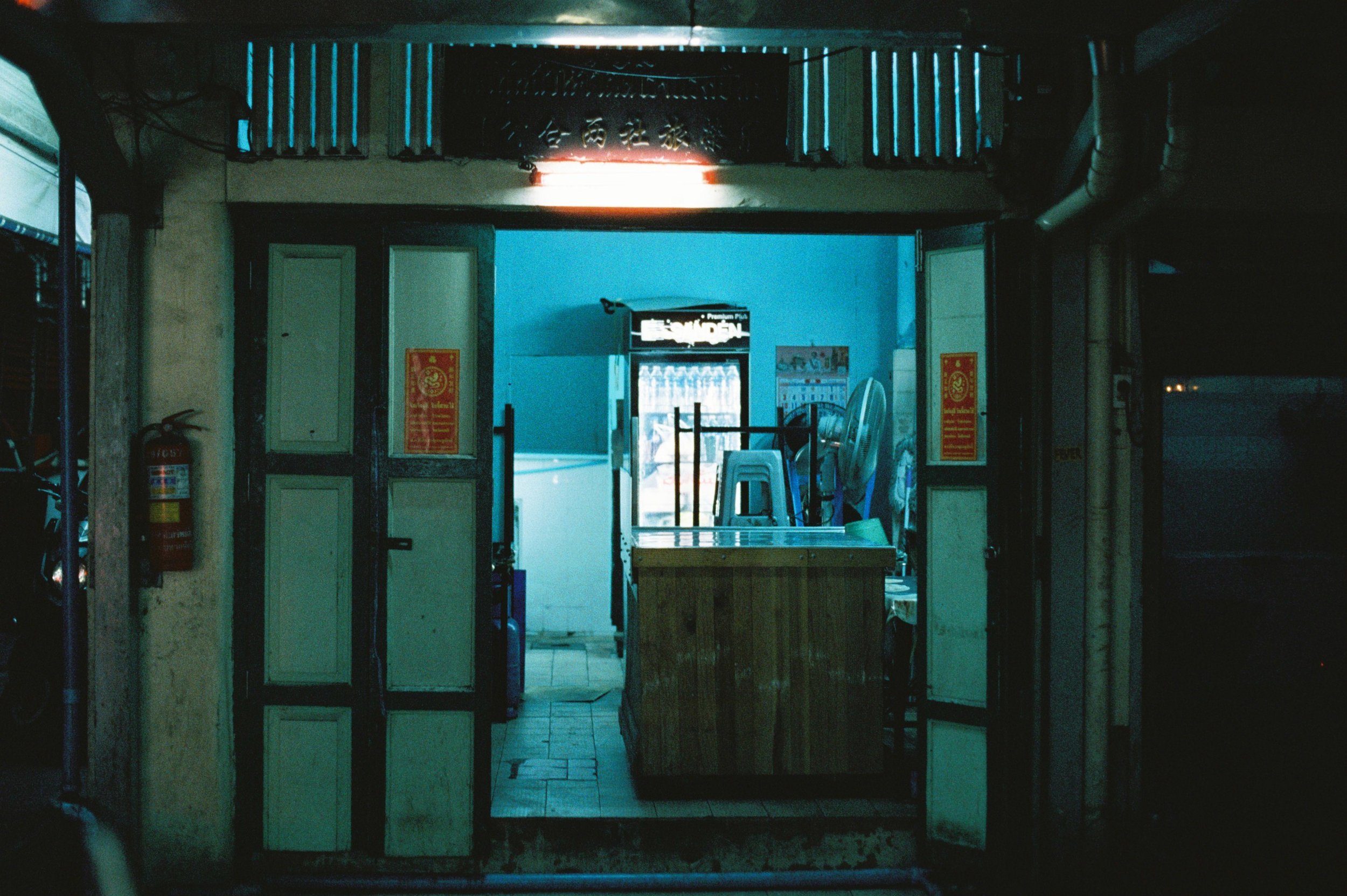 Vista de un negocio cerrado con puertas abiertas, con sillones apilados y una mesa en el interior, ambiente iluminado tenuemente con luz azul, y un extintor en la pared izquierda.