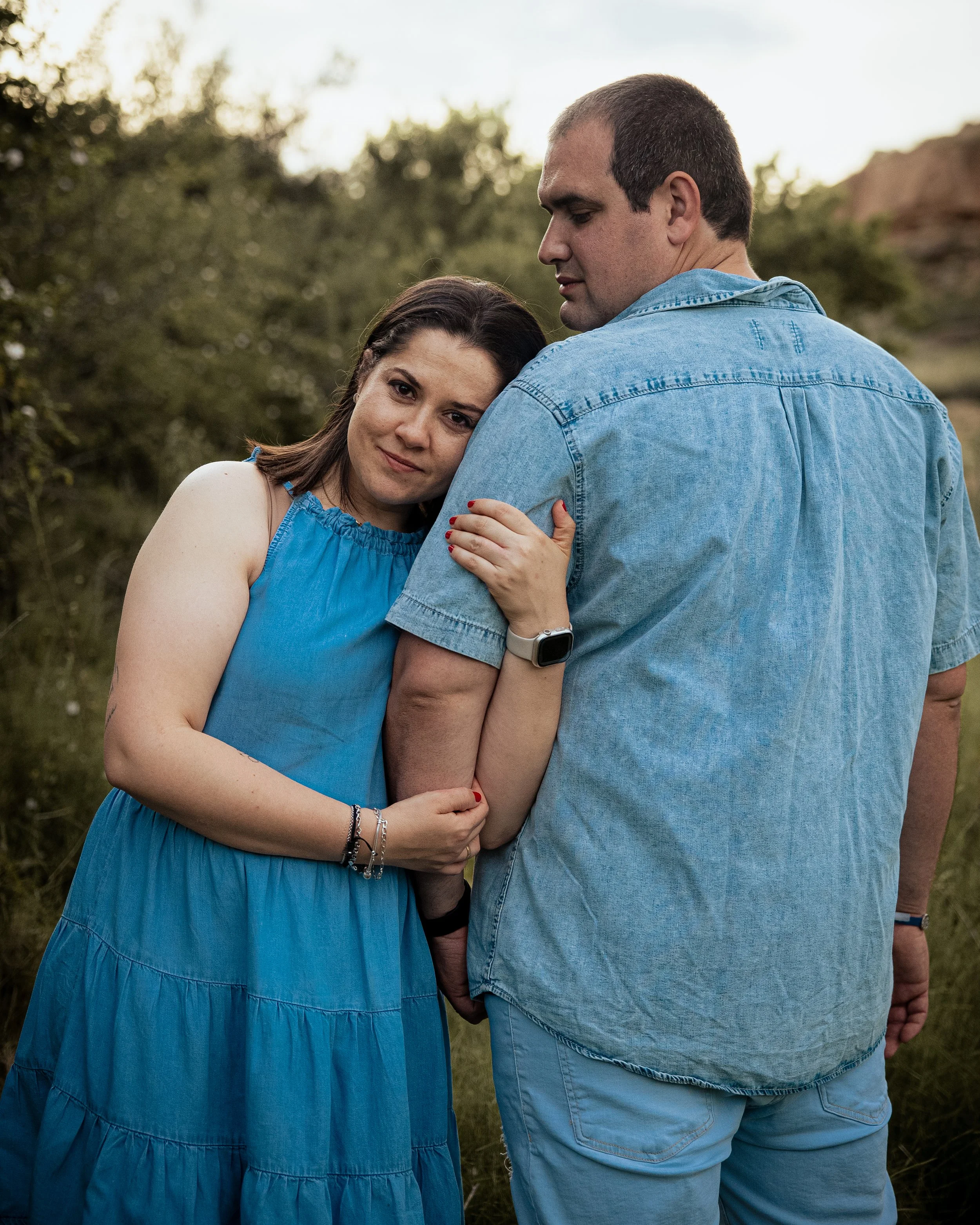 Una pareja en un campo, la mujer abraza al hombre y se apoya en su hombro, una escena al atardecer.