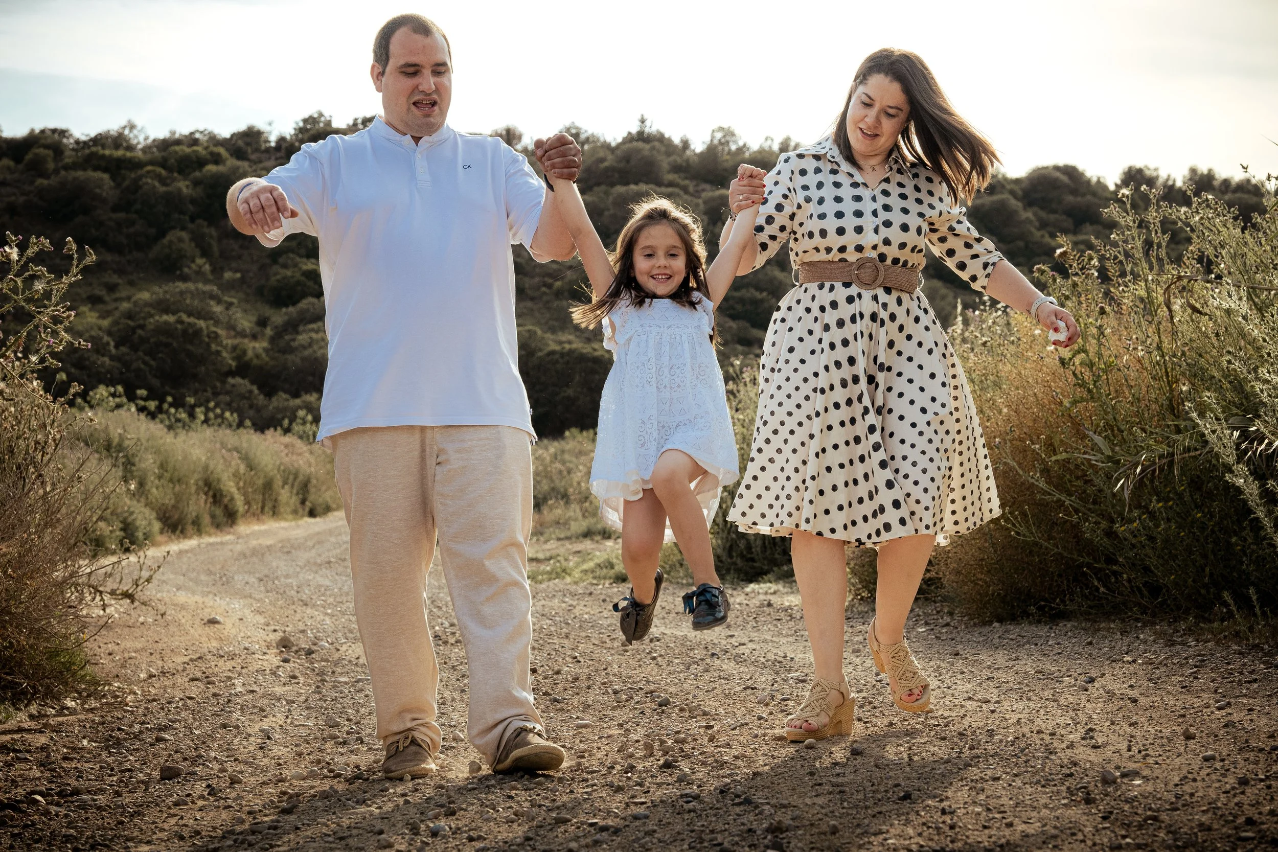 Familia de cuatro caminando en un sendero en la naturaleza, sosteniendo las manos de una niña pequeña que está siendo levantada en el aire.