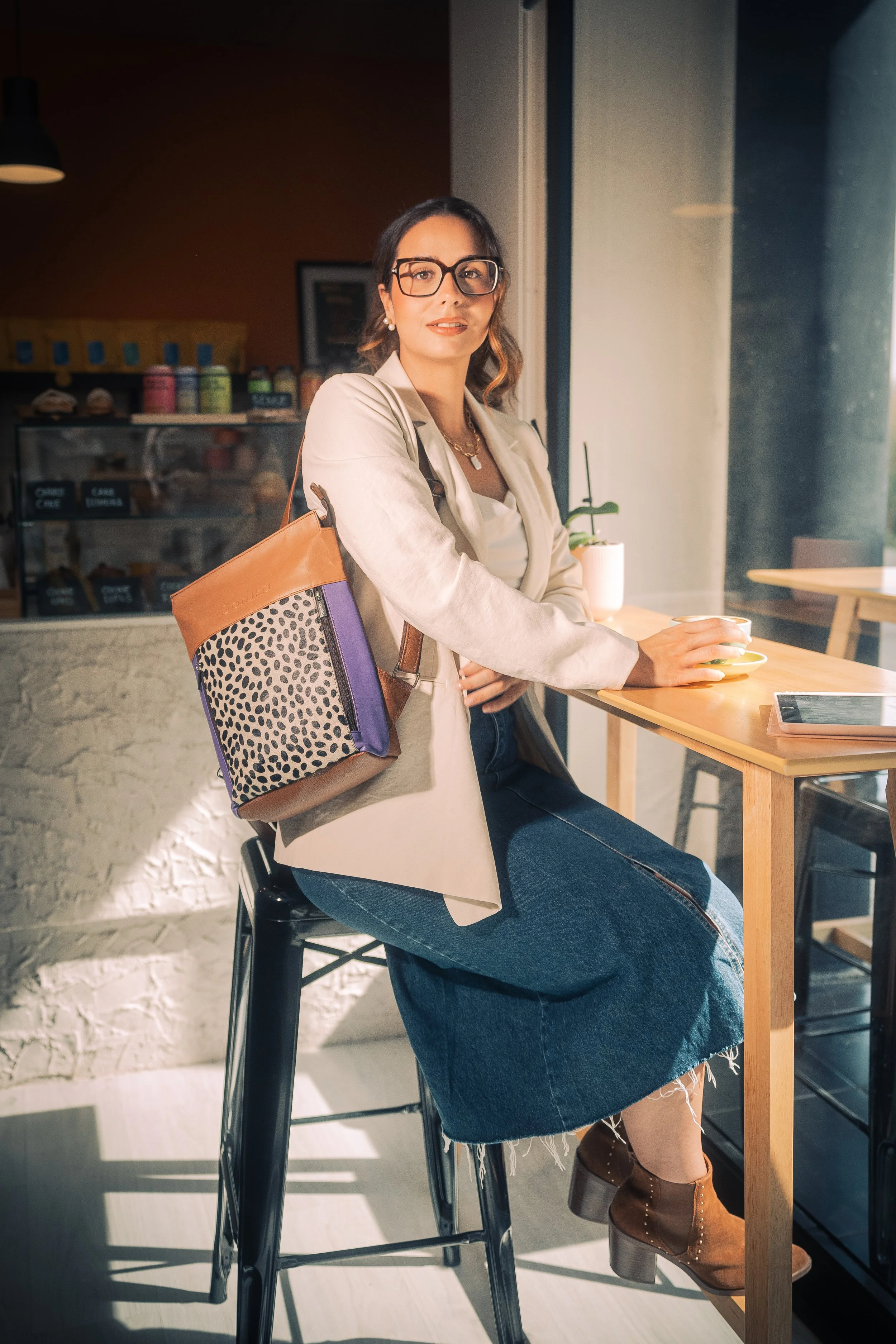 Mujer sentada en una barra de café con gafas, collar y bolso de mano, sosteniendo una taza de café en una mano y mirando hacia la cámara.