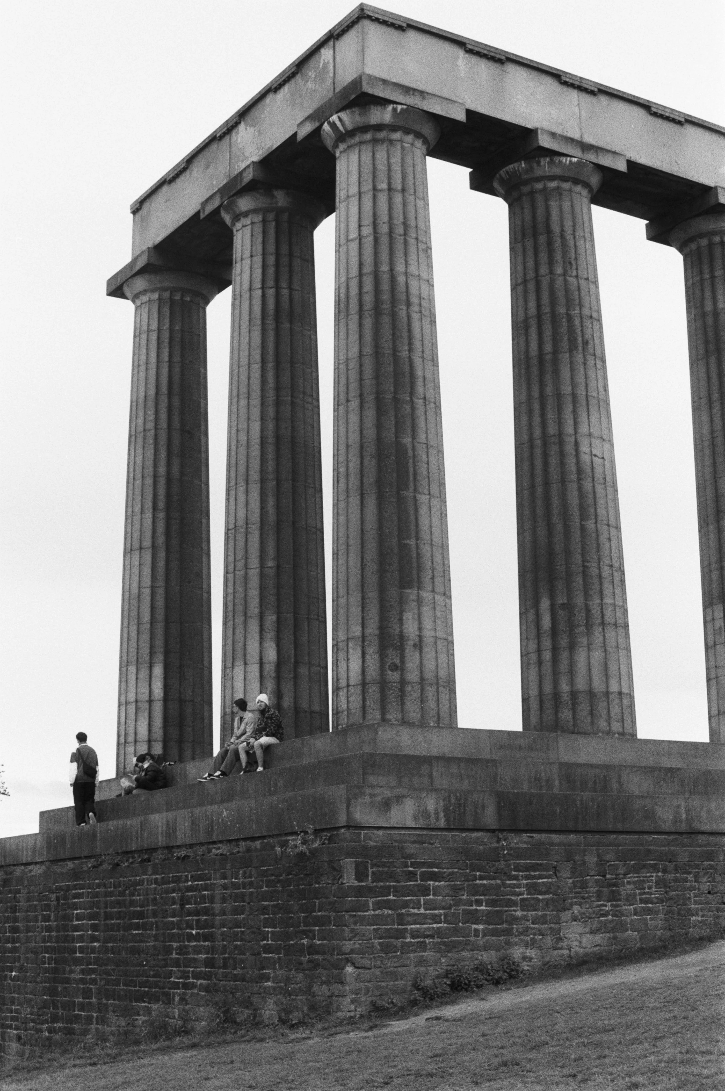 Vista en blanco y negro de la estructura de un templo con columnas grandes y altas, con varias personas sentadas y de pie en la base de las columnas.