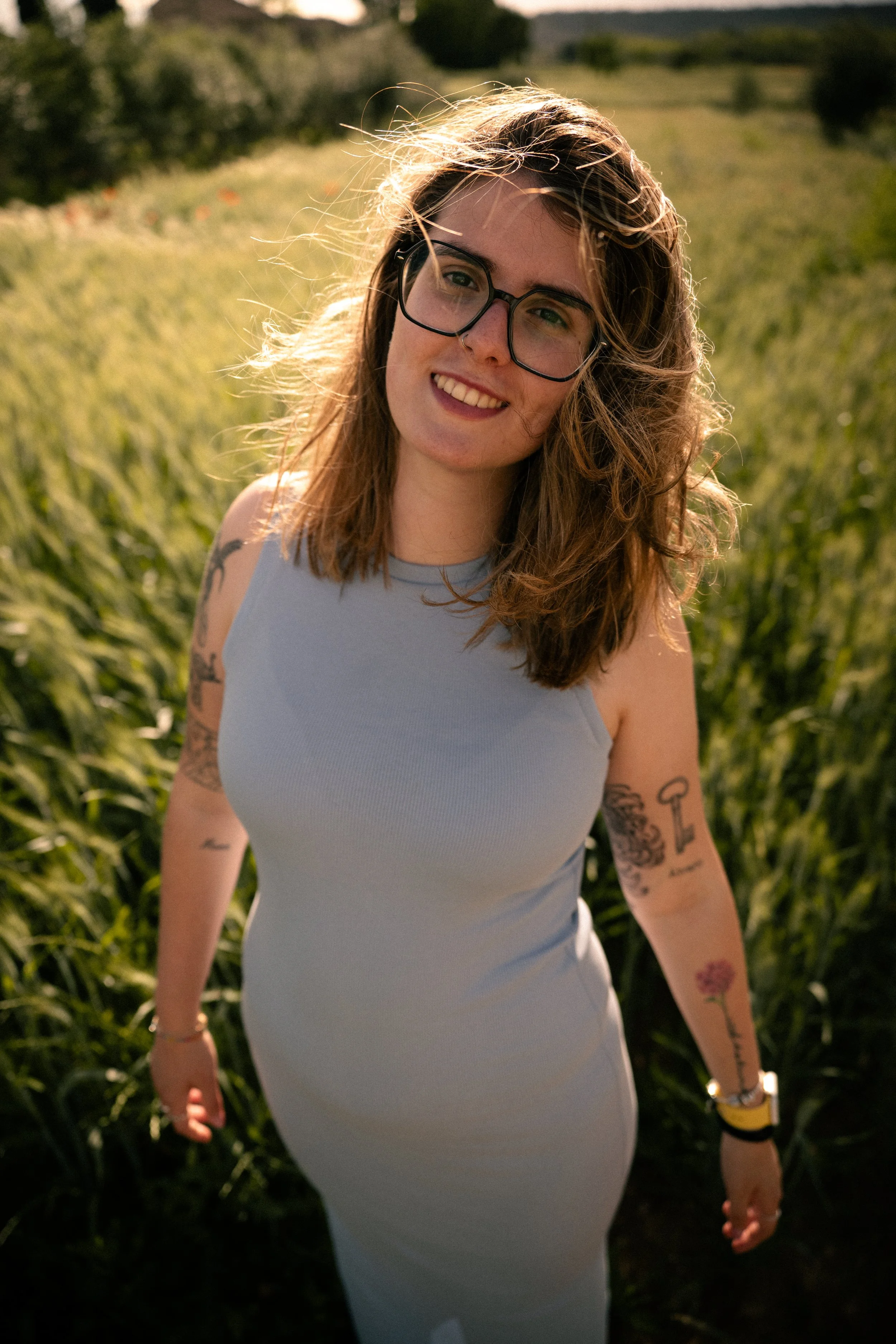 Mujer con gafas y tatuajes en un campo verde al atardecer.