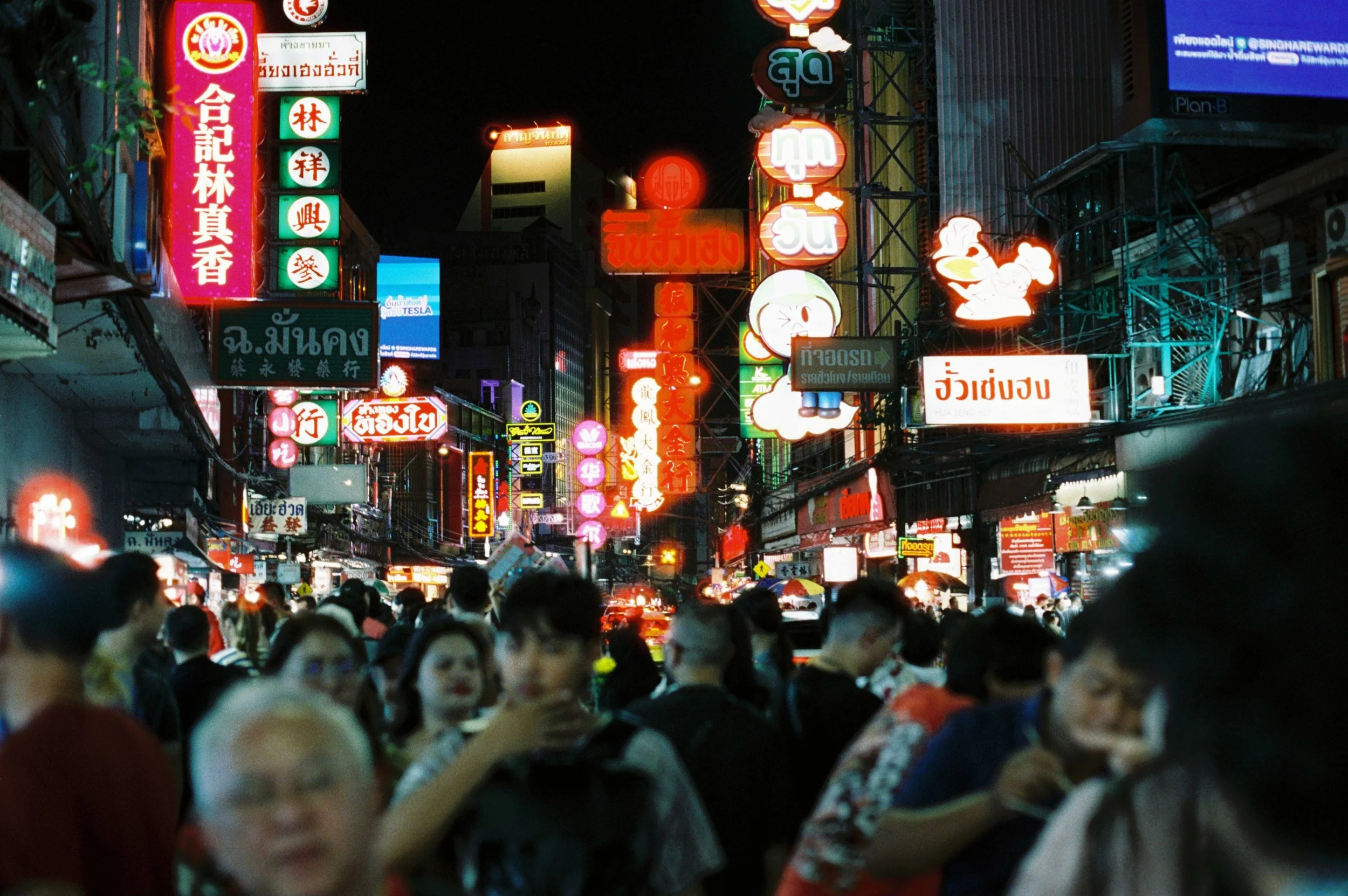 Una calle concurrida en un área urbana con carteles de neón en idioma tailandés iluminados por la noche, y muchas personas caminando en la calle.