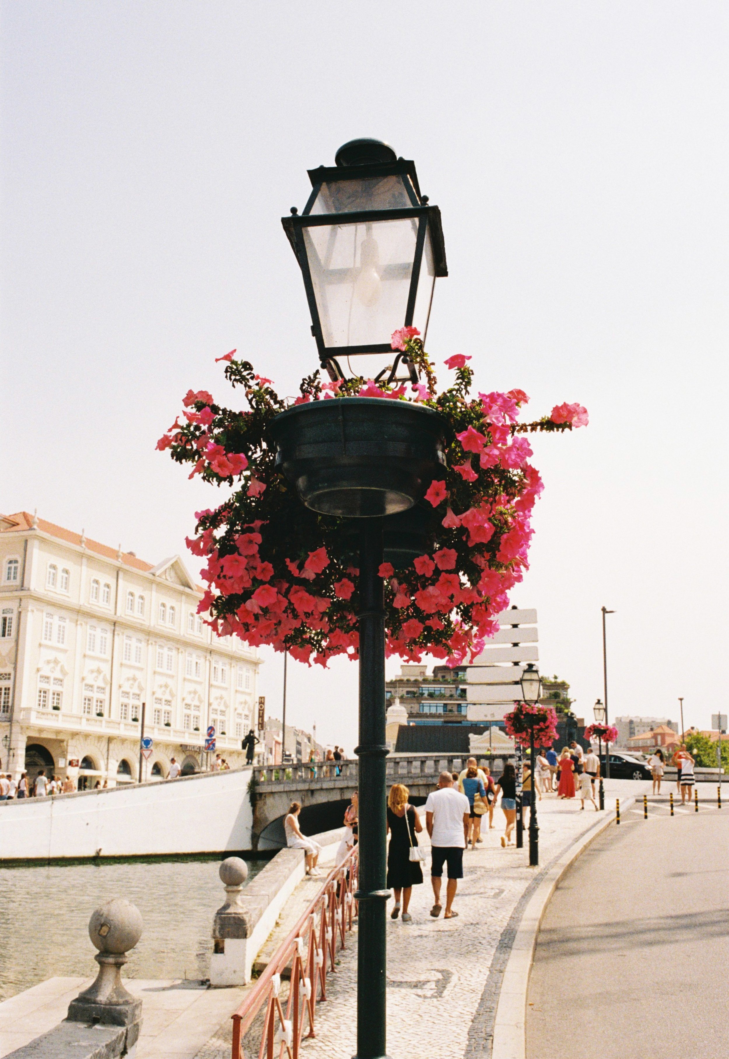 Farola decorada con flores rosas en una acera junto a un canal y un puente en una ciudad animada con gente caminando y edificios en el fondo.