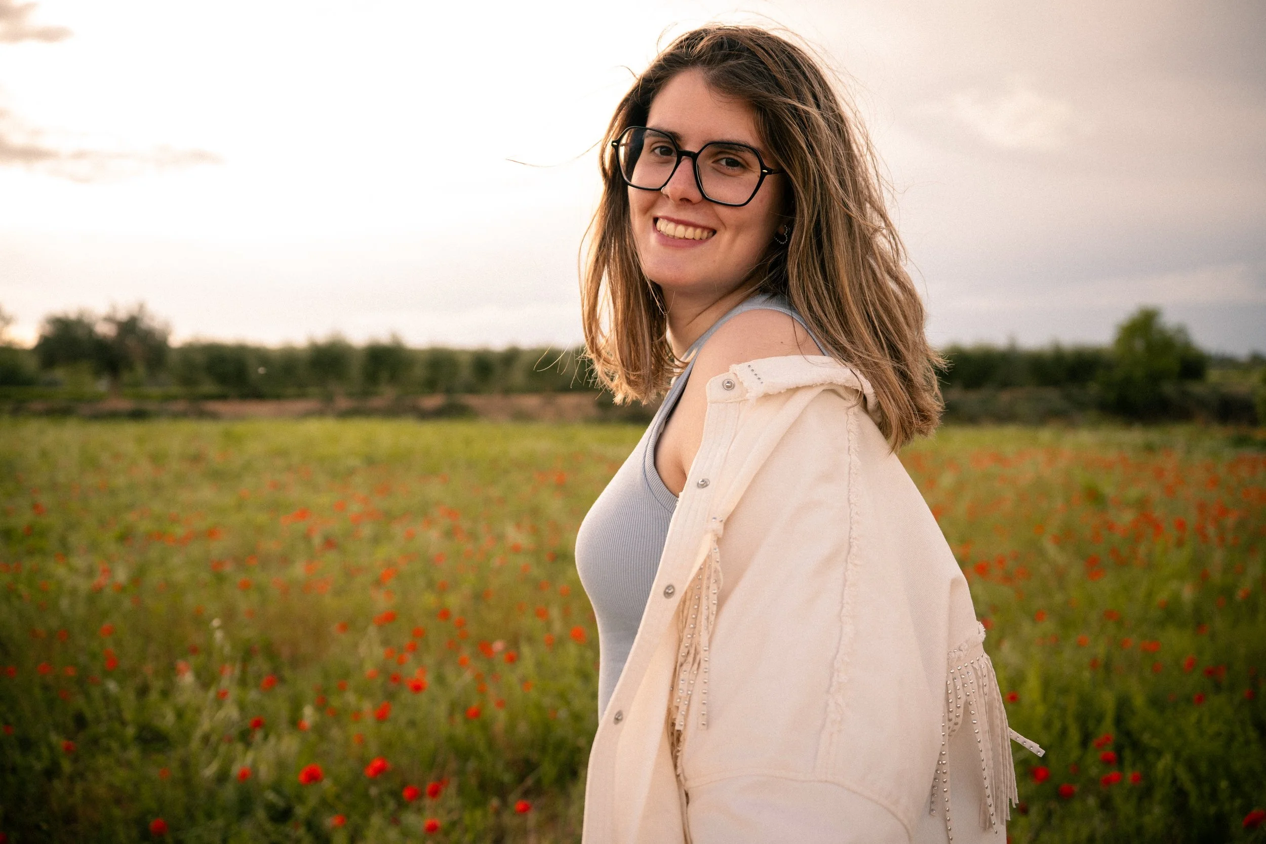 Joven mujer sonriendo en un campo con flores rojas, viste una camiseta gris y una chaqueta beige, lleva gafas y tiene el cabello castaño medio.