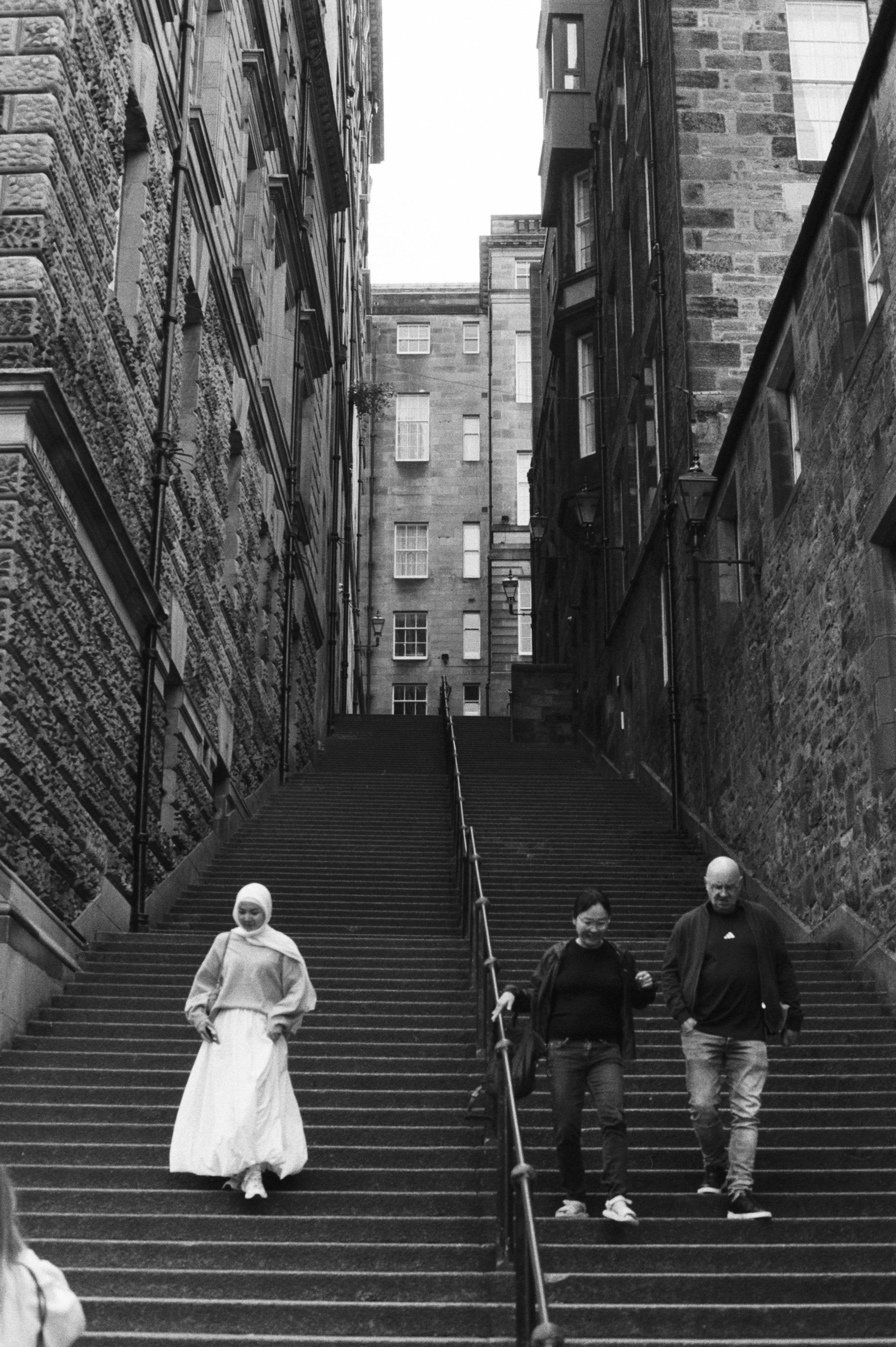 Tres personas bajando unas escaleras en una calle urbana con edificios de piedra a ambos lados, en un estilo en blanco y negro.
