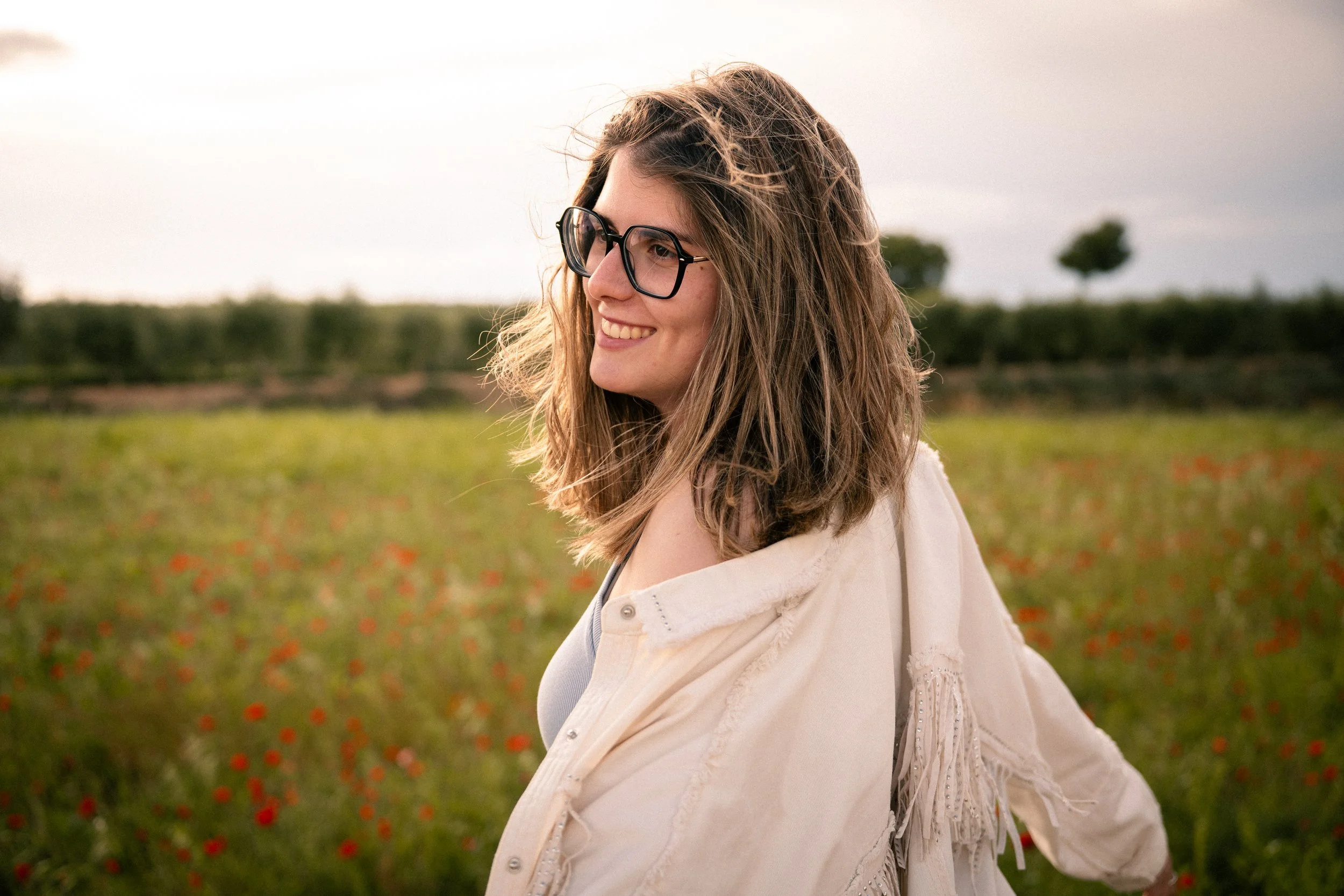 Joven mujer con gafas, sonriendo en un campo con flores, vestido blanco y una chaqueta