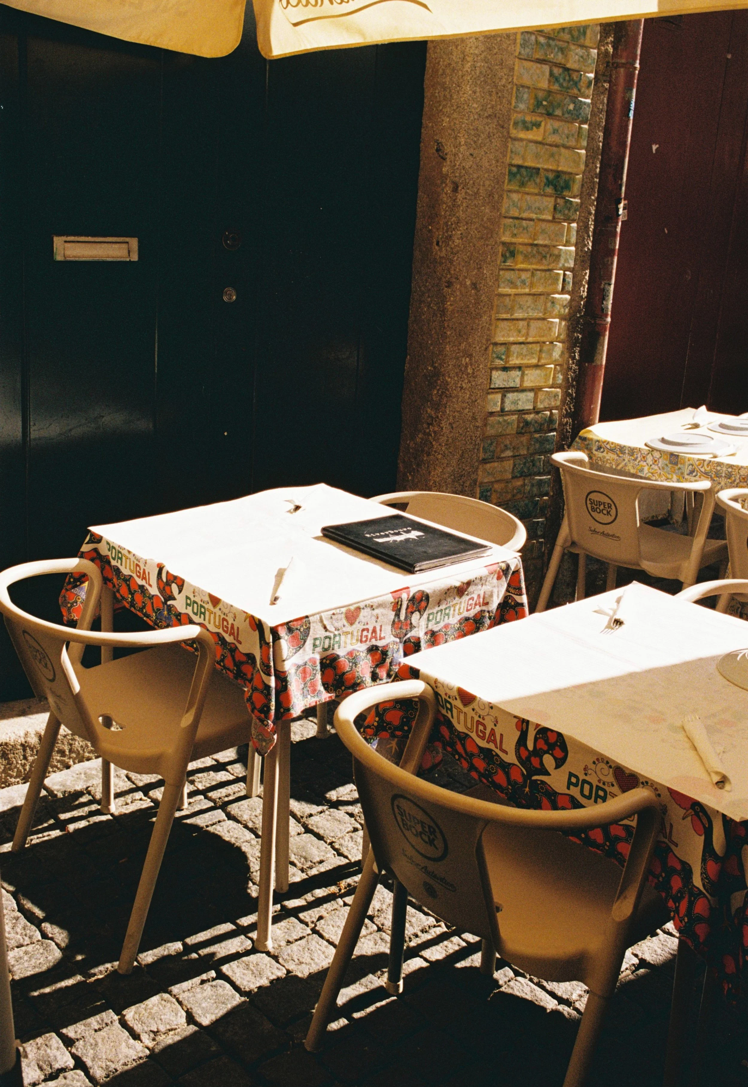Mesas de restaurante al aire libre con manteles decorados con motivos portugueses y sillas blancas, situadas sobre calles de piedra. Se observa un menú en una de las mesas y un entorno con paredes de ladrillo y colores cálidos.