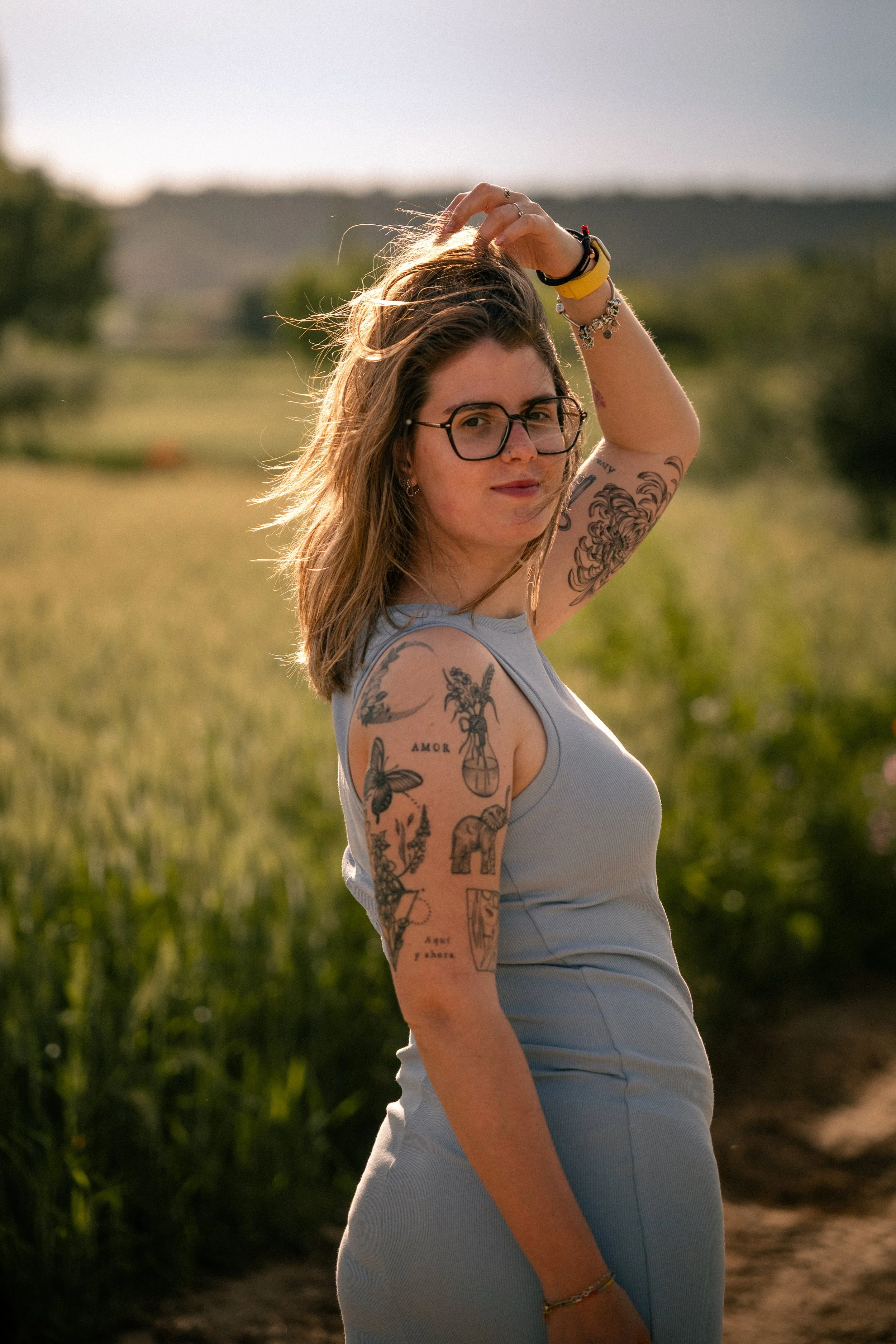 Joven mujer con gafas, cabello castaño, tatuajes en los brazos, usando un vestido sin mangas en campo al atardecer.