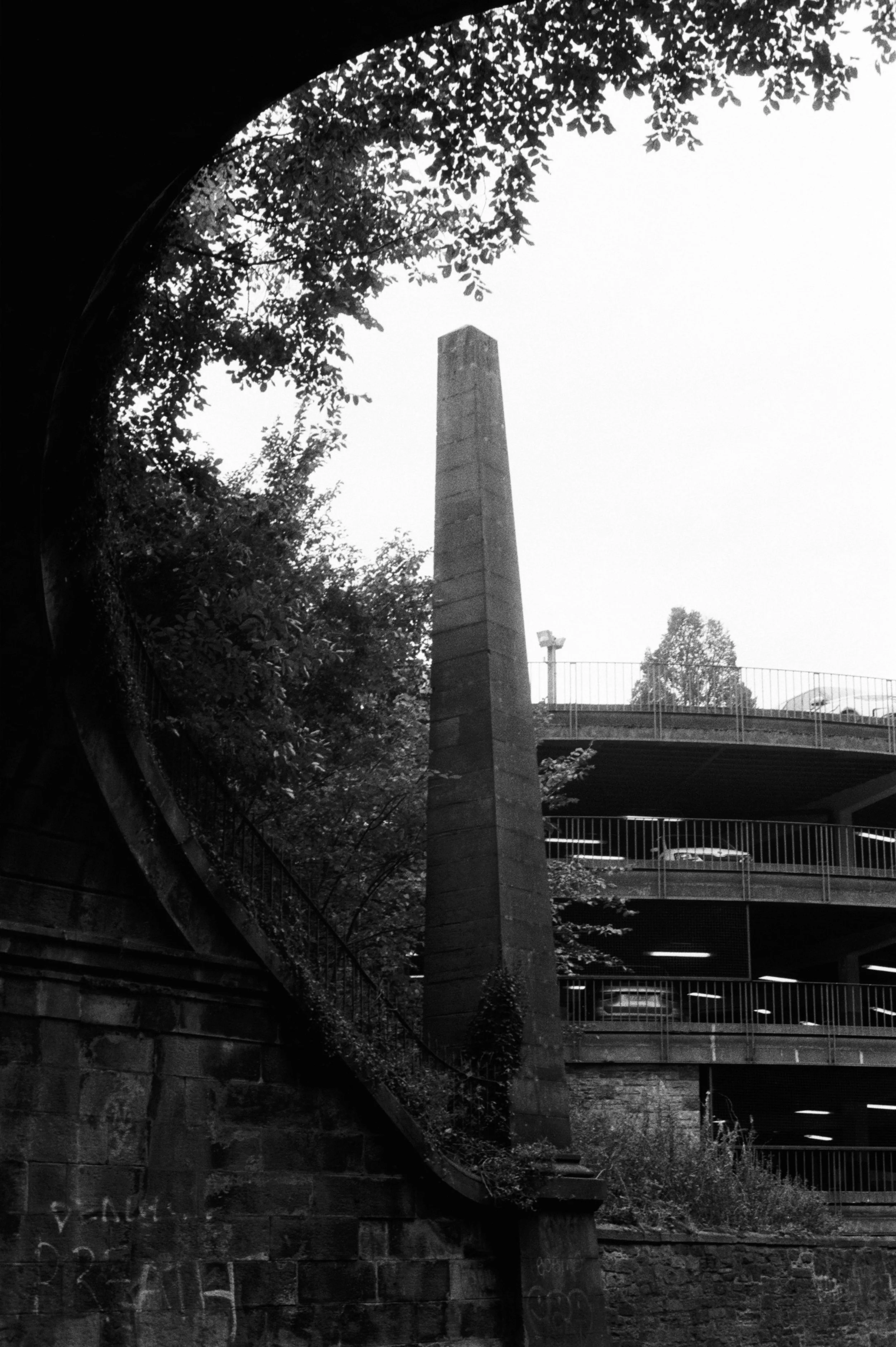 Estructura de un estacionamiento con varios niveles y un muro de ladrillos, en primer plano hay un alto pilón o columna de piedra, rodeada de árboles y vegetación.