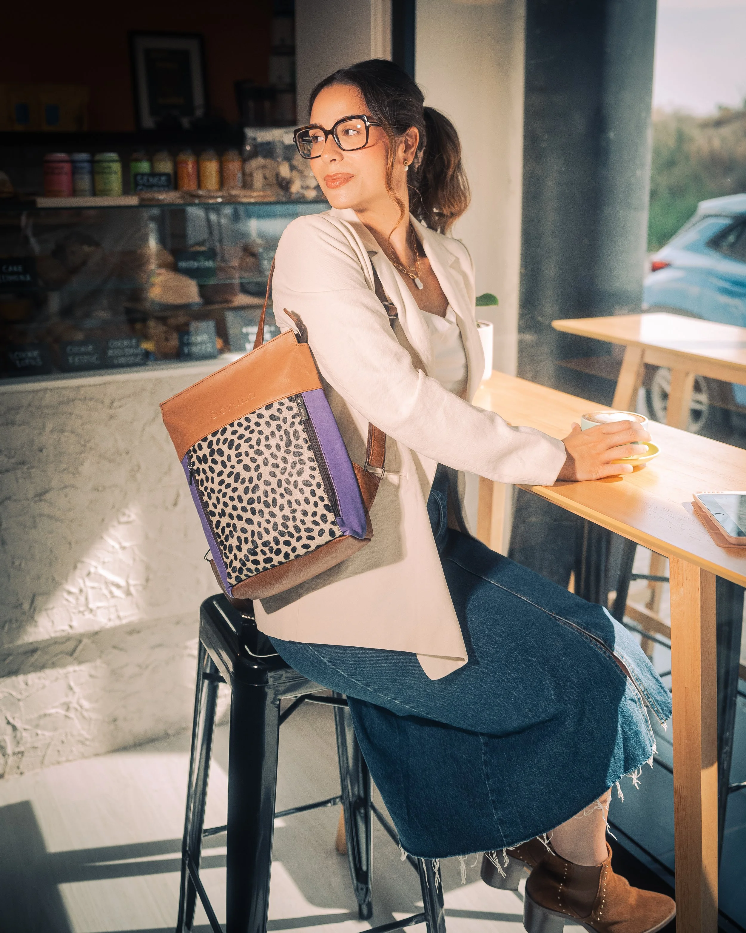 Mujer con gafas, ropa beige y falda de mezclilla, sentada en una mesa de cafetería con una taza en mano, en un ambiente soleado con vista a la calle.
