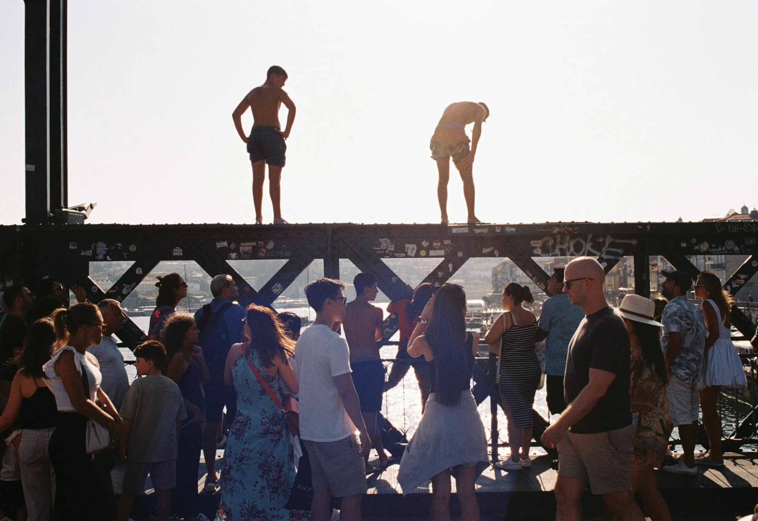 Personas y jóvenes en un puente peatonal con vista al río, con dos niños en la parte superior del puente en silhouetta al sol, mientras otras personas caminan y conversan en la parte inferior.