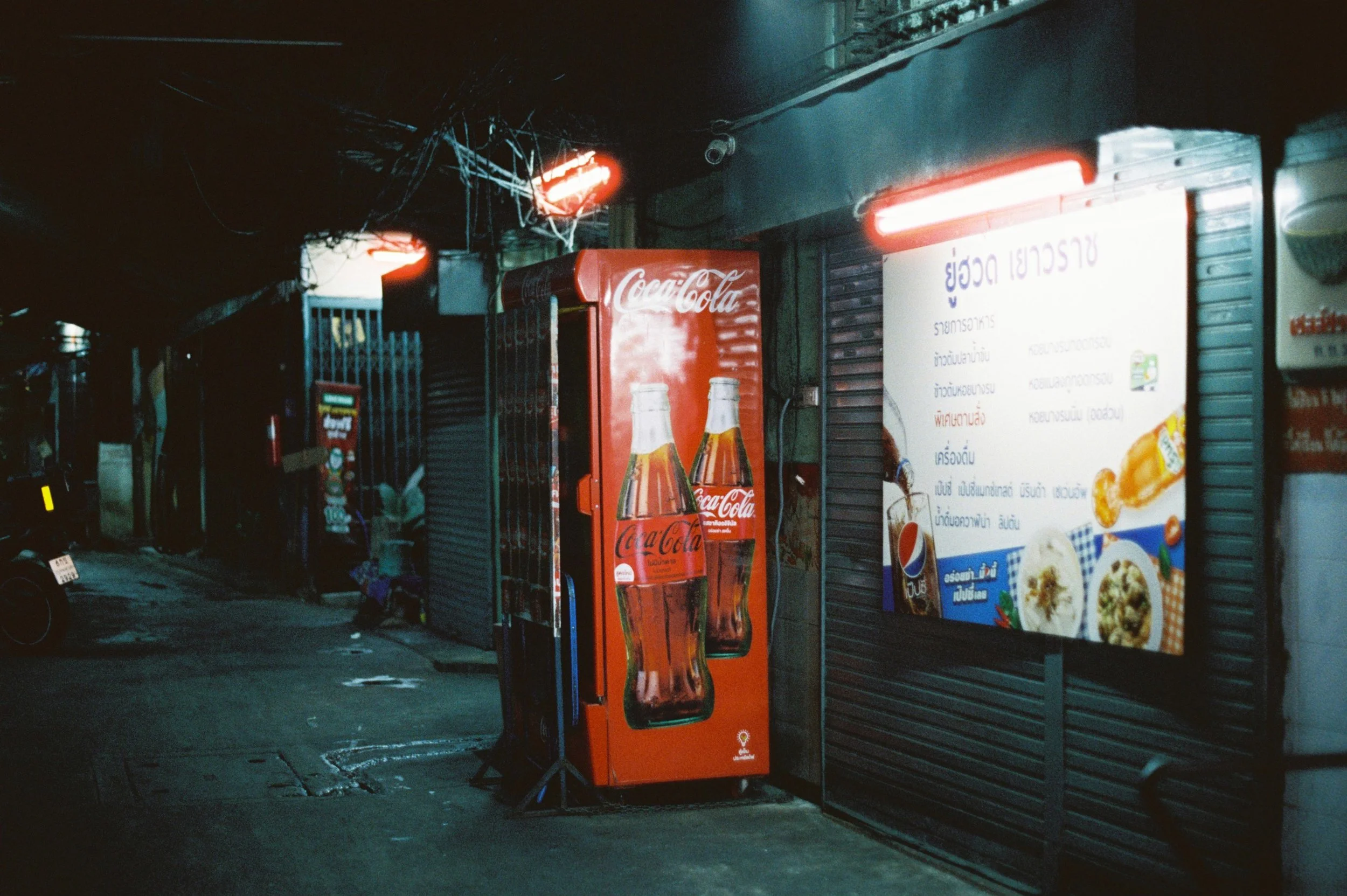 Escena nocturna en una calle de Tailandia con carteles en tailandés y una máquina expendedora de Coca-Cola con botellas de vidrio y un cartel de menú con comida tailandesa.