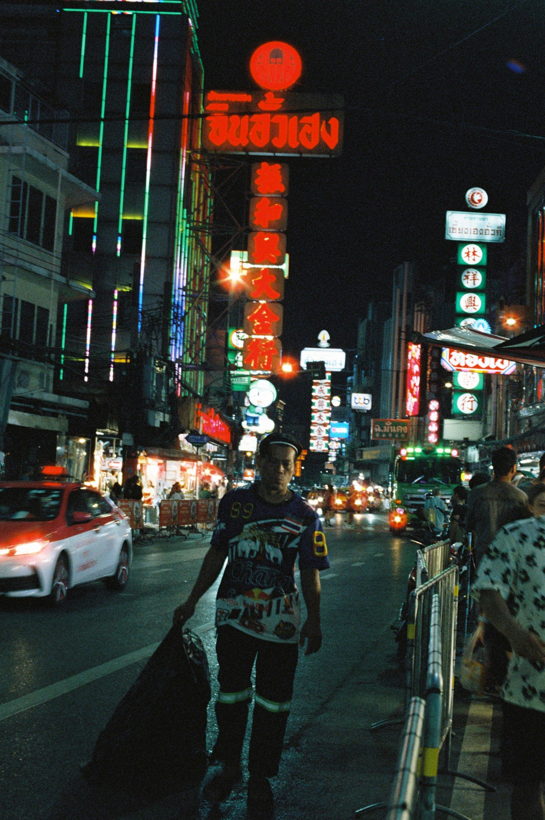 Una calle nocturna en una ciudad con signos en chino y luces de neón, varias personas caminando y autos pasando, con un hombre en el primer plano que lleva una bolsa y ropa deportiva.