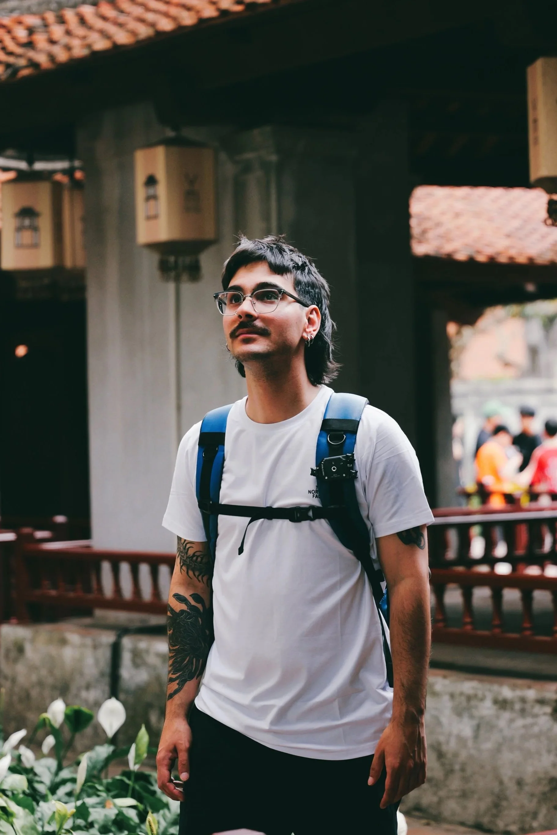 Joven con gafas, barba y tatuajes en brazos, lleva mochila y camiseta blanca, en un entorno al aire libre con fondo de casas y flores.