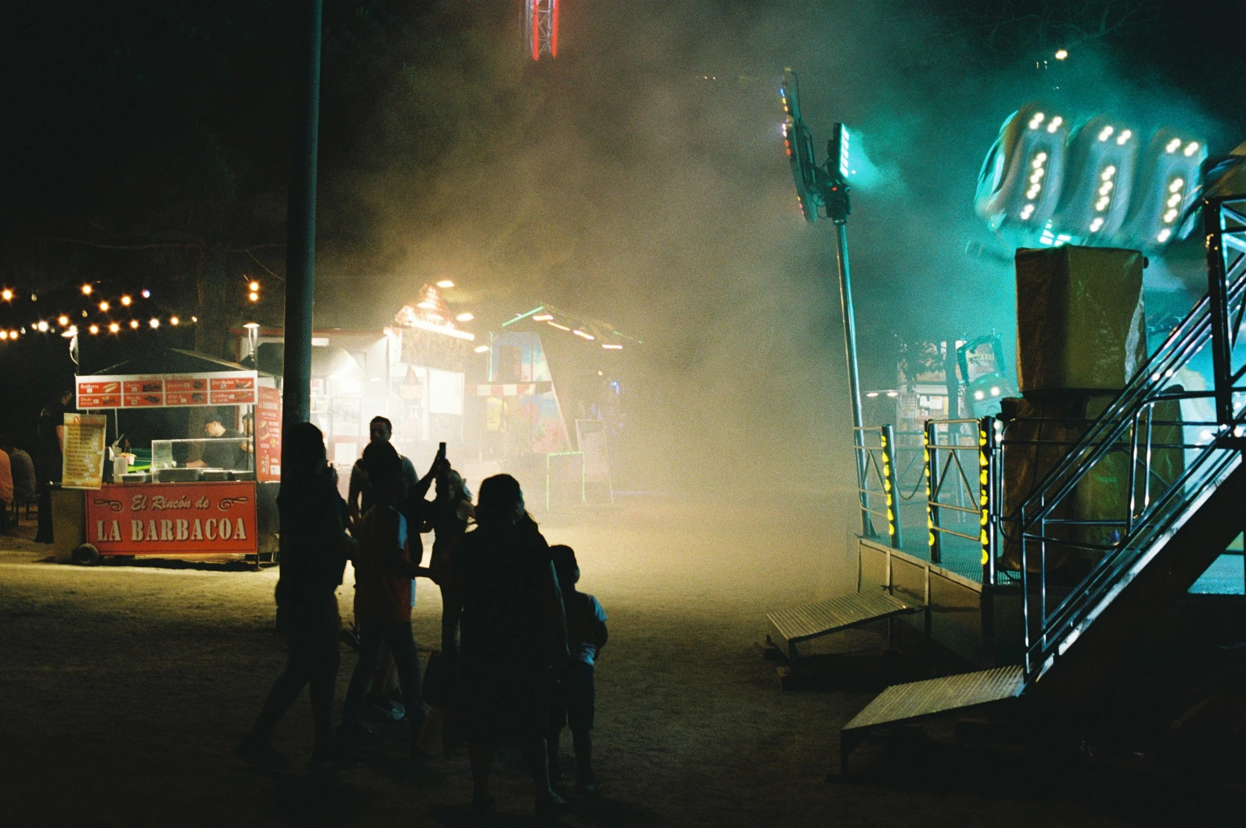 Personas observando una atracción en un parque de diversiones durante la noche, con luces y estructuras de rides en el fondo.