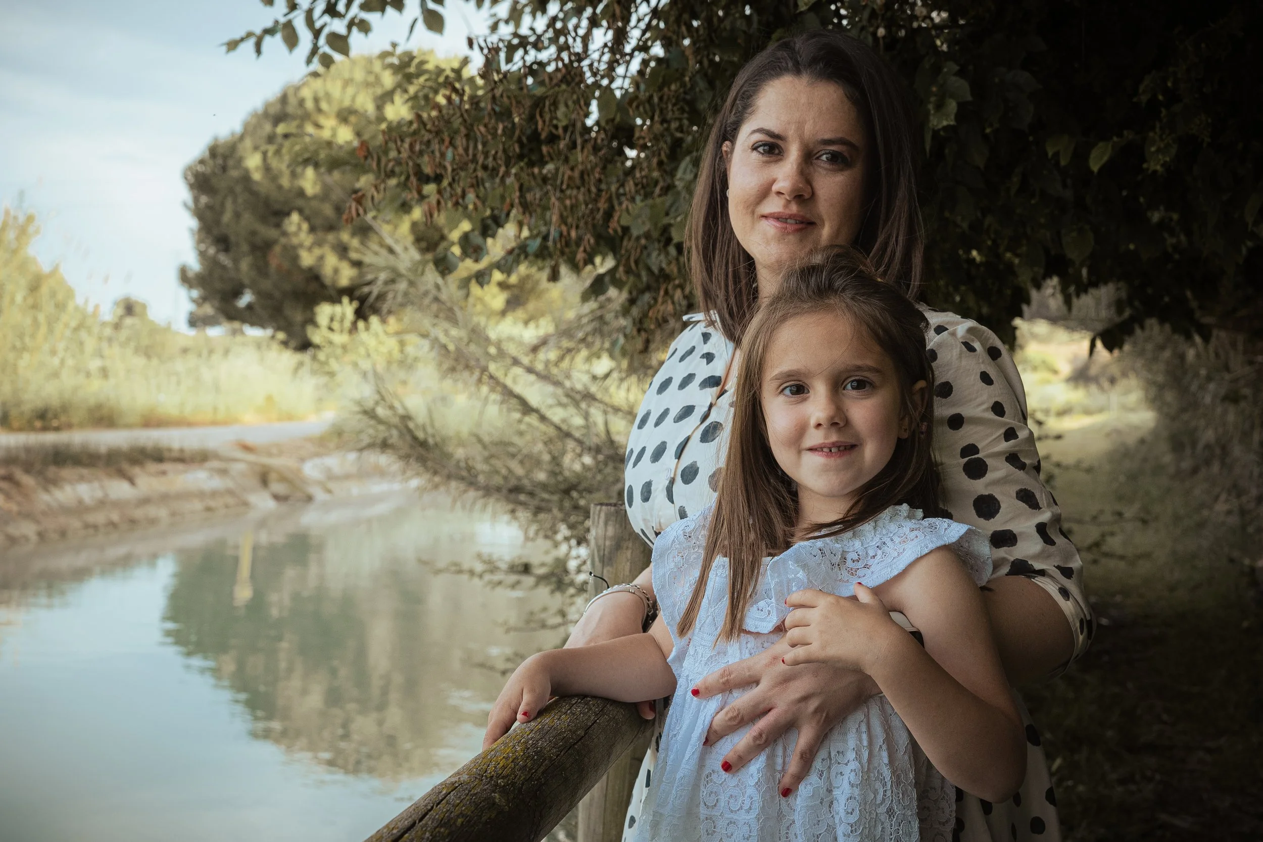 Una mujer y una niña posando juntas junto a un río, rodeadas de árboles y vegetación.