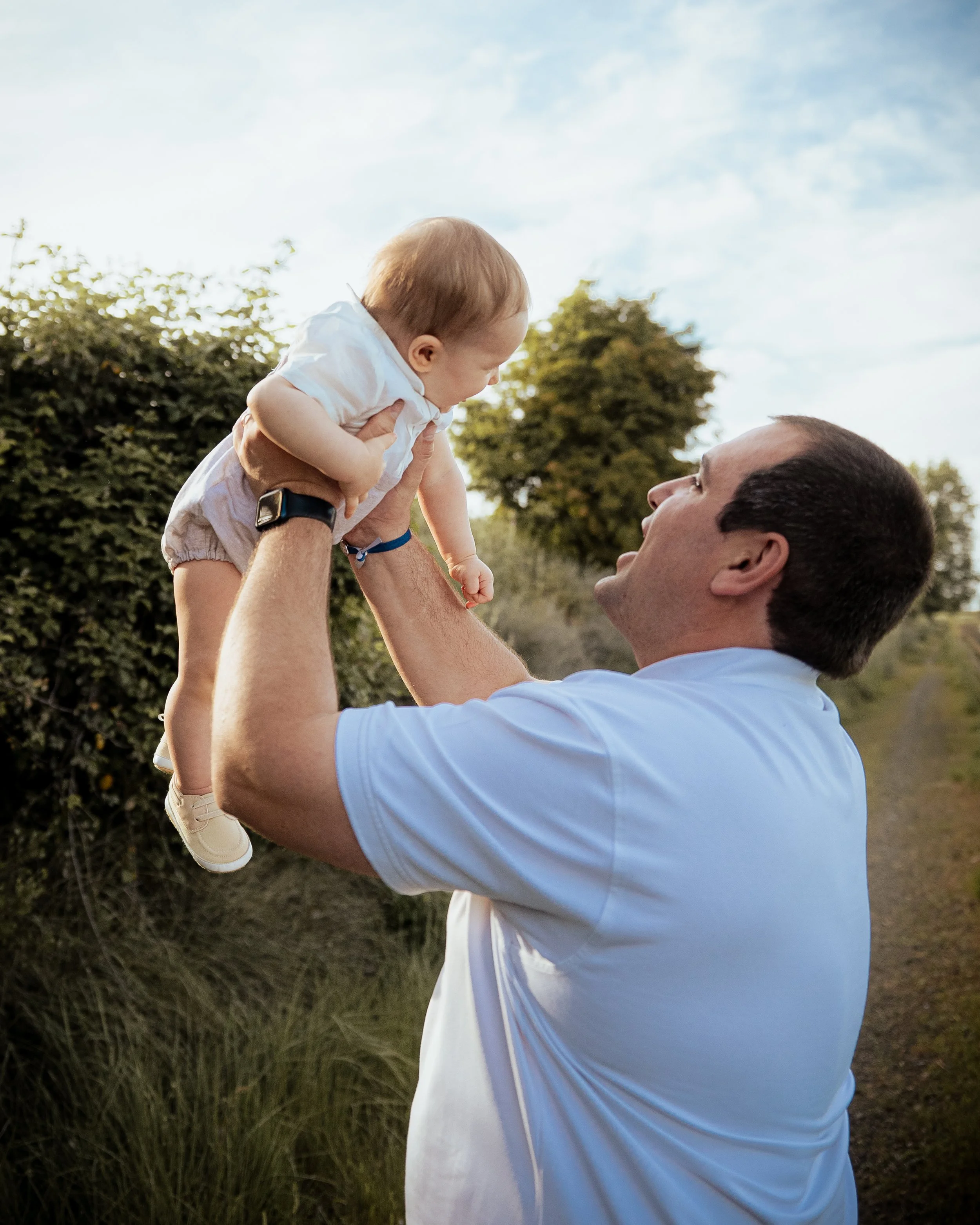 Un hombre levantando a un bebé en un ambiente al aire libre con árboles y cielo azul