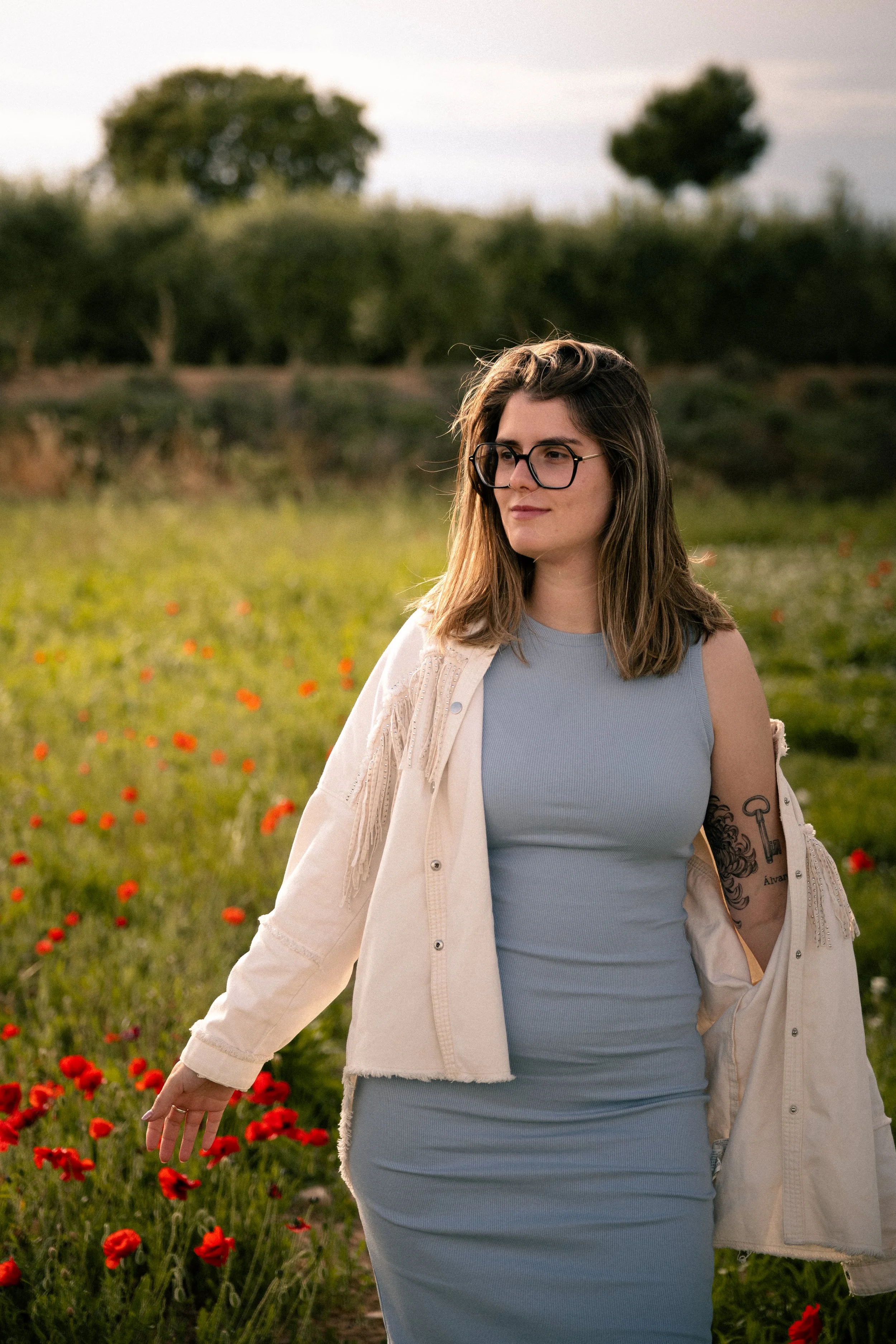 Mujer de cabello castaño, gafas y tatuajes, caminando por un campo con flores rojas en un atardecer o anochecer.
