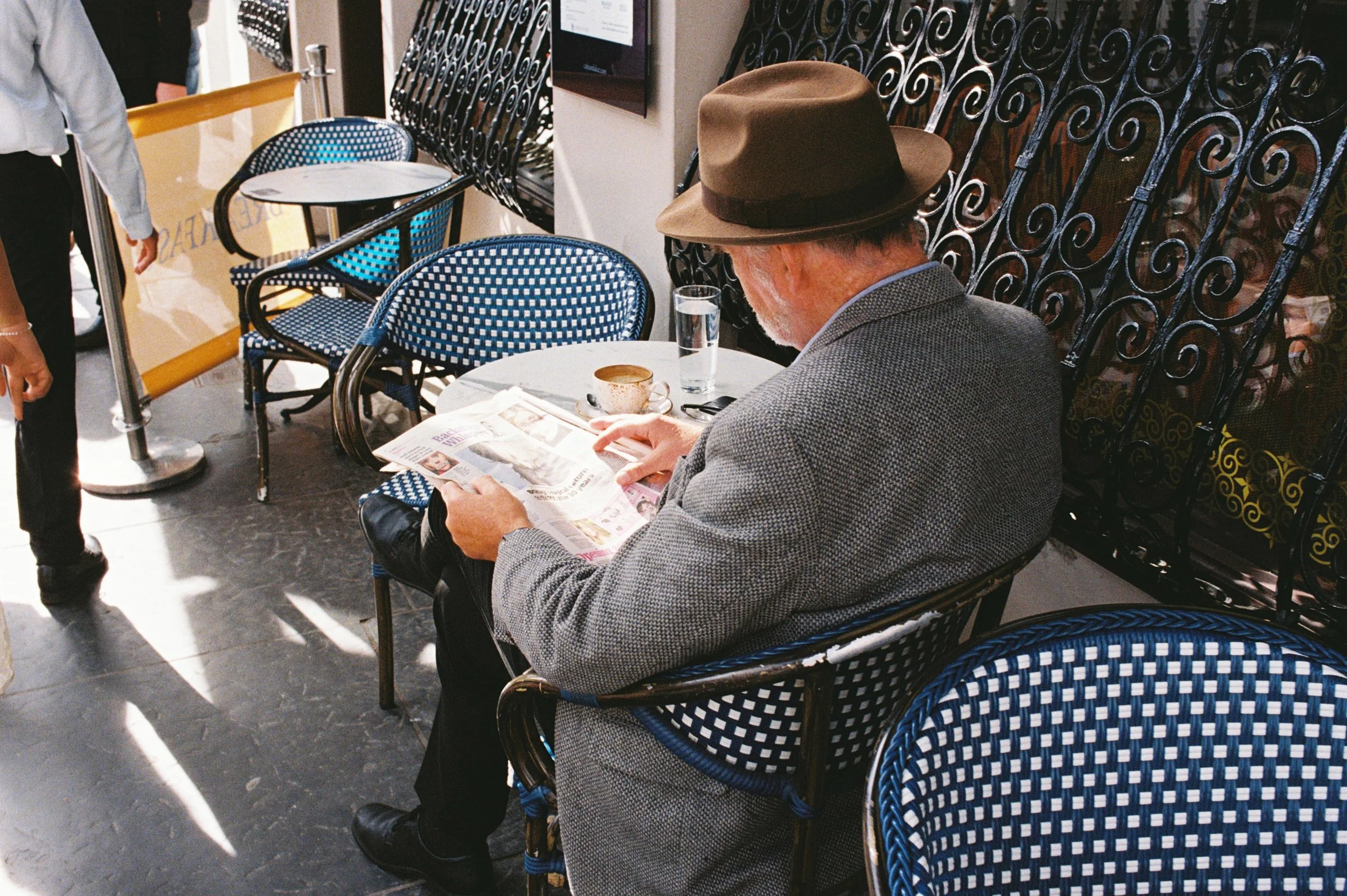 Hombre mayor con sombrero y saco gris leyendo el periódico en una cafetería, acompañado de una taza y un vaso de agua sobre la mesa.