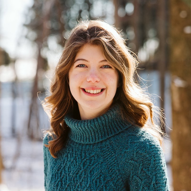 A woman with shoulder-length light brown hair wearing a teal turtleneck sweater, smiling outdoors on a sunny winter day with blurred trees in the background.