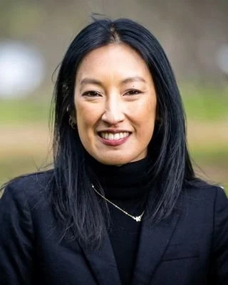 Portrait of a smiling Asian woman with long black hair wearing a black blazer and a gold necklace, outdoors with blurred background.