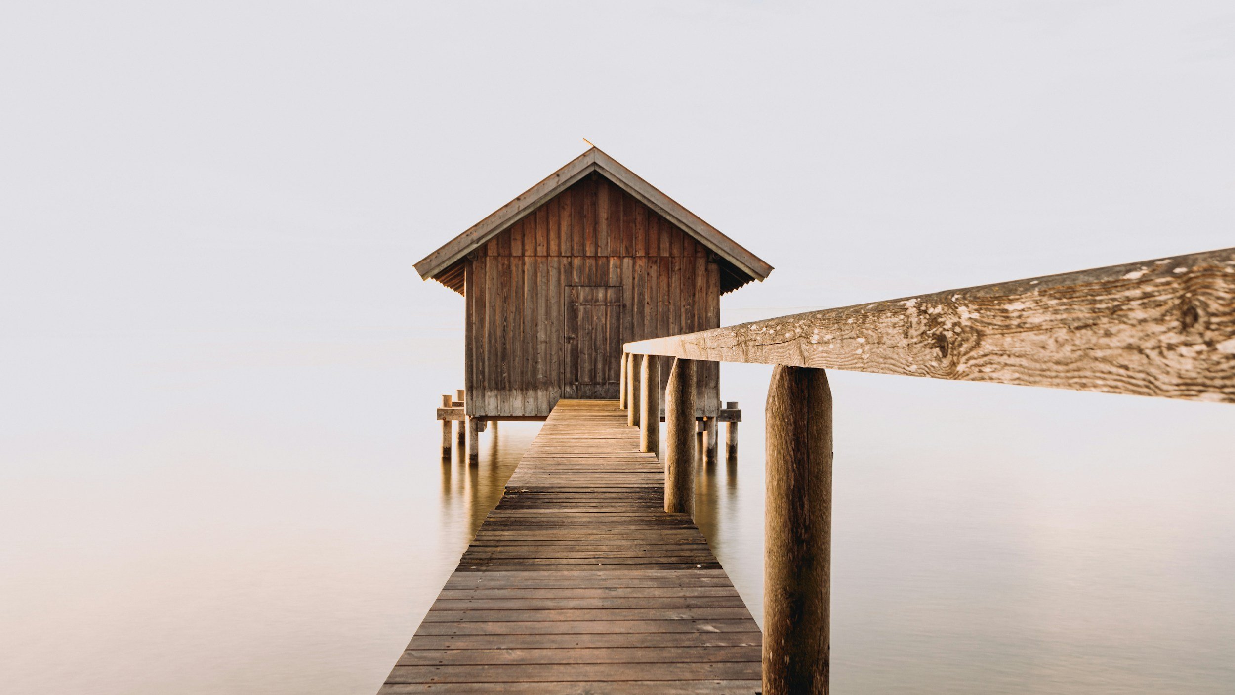 A wooden dock extends into calm water, leading to a small wooden cabin on stilts with a pitched roof, under a clear sky.