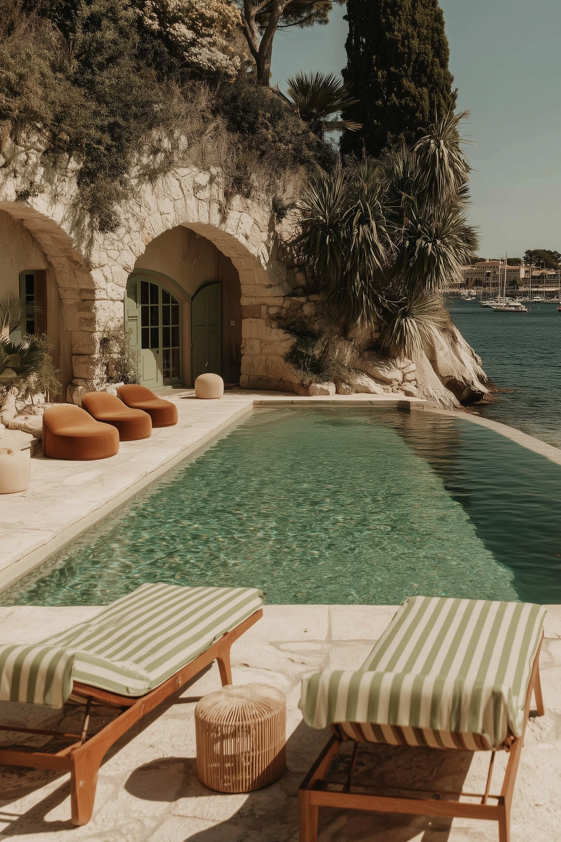 Luxury seaside pool area with two striped lounge chairs, a small round wicker table, three chaise lounges, lush greenery on a rocky hillside, and boats docked in a marina in the background.