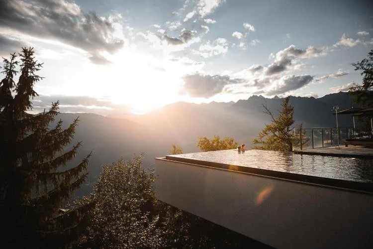 Sunset viewed from a modern building with an infinity pool, overlooking mountains and trees.