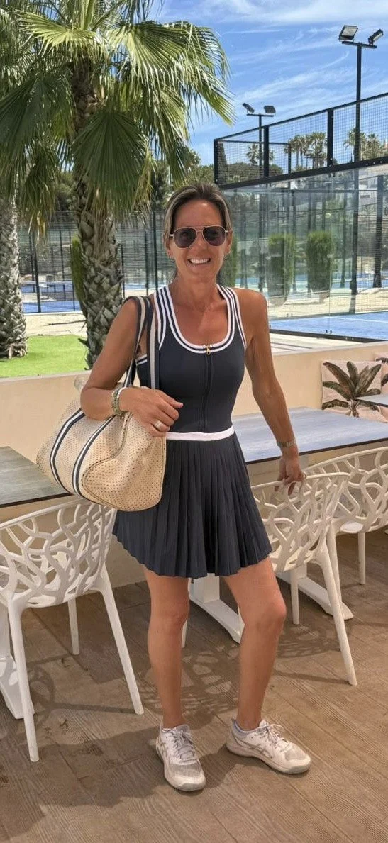 A woman with short brown hair wearing sunglasses, a sleeveless black sports top, a black pleated skirt, and white sneakers, standing outdoors near tennis courts, holding a large beige handbag, smiling, with palm trees and a blue sky in the background.
