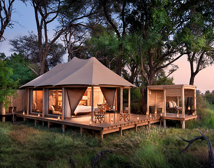 Luxury tent and outdoor lounge areas on a wooden deck surrounded by trees at sunset.