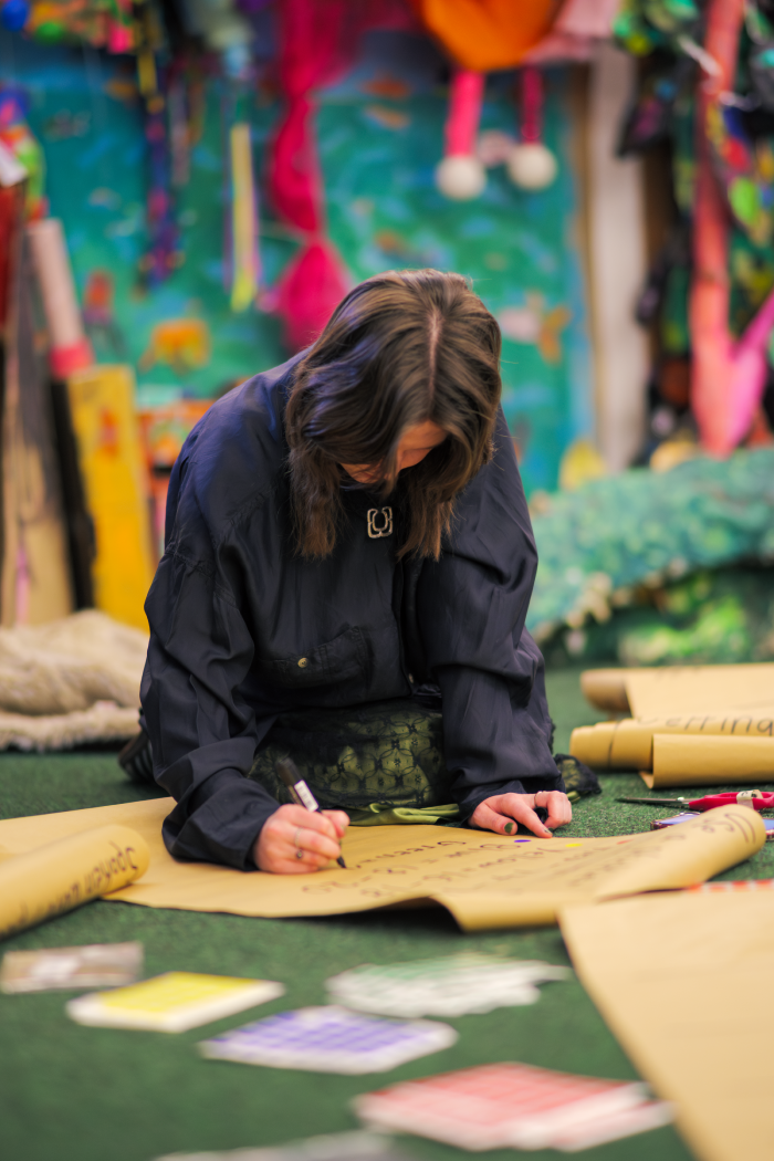A woman with shoulder-length brown hair, dressed in a black shirt, is kneeling on the floor, writing on a large paper with a black marker. The setting appears to be an art or creative space with colorful decorations and supplies in the background.