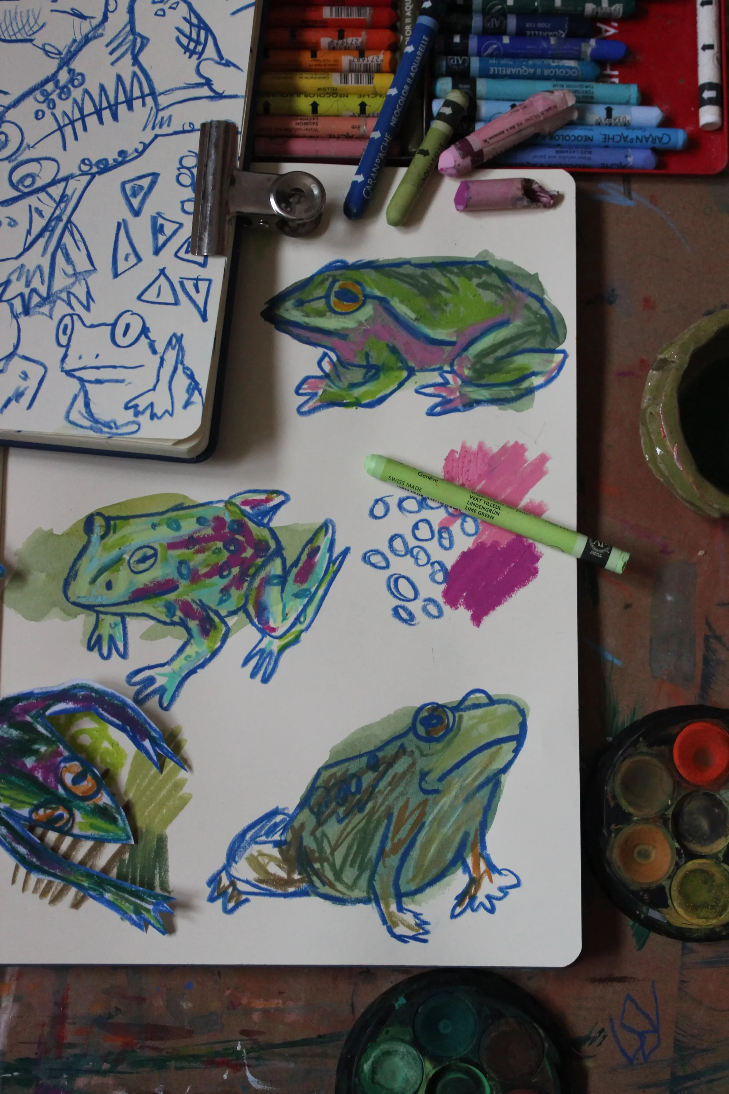 Colorful sketches of frogs and reptiles created with markers, including some colored in green, pink, and purple, on paper. Markers and paint containers are also visible on the table.