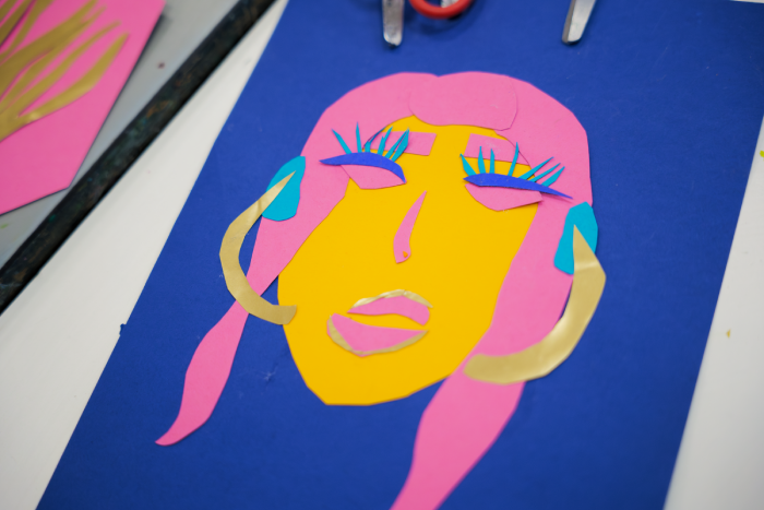 Colorful paper collage of a woman's face with pink hair, blue eyelashes, and large earrings on a blue background. Created at a workshop.
