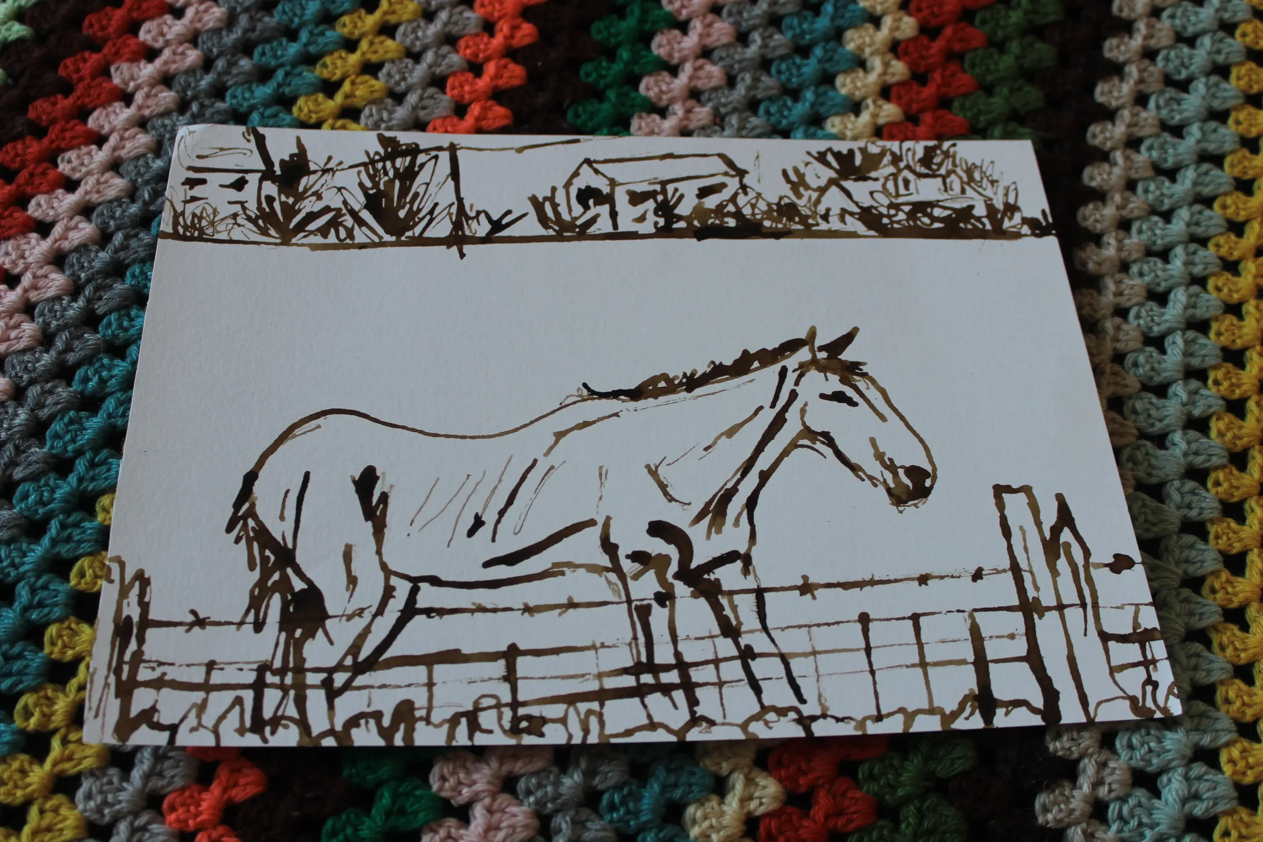 A white card with a brown ink sketch of a rural landscape featuring a house, trees, and a horse on a multicoloured crocheted blanket.