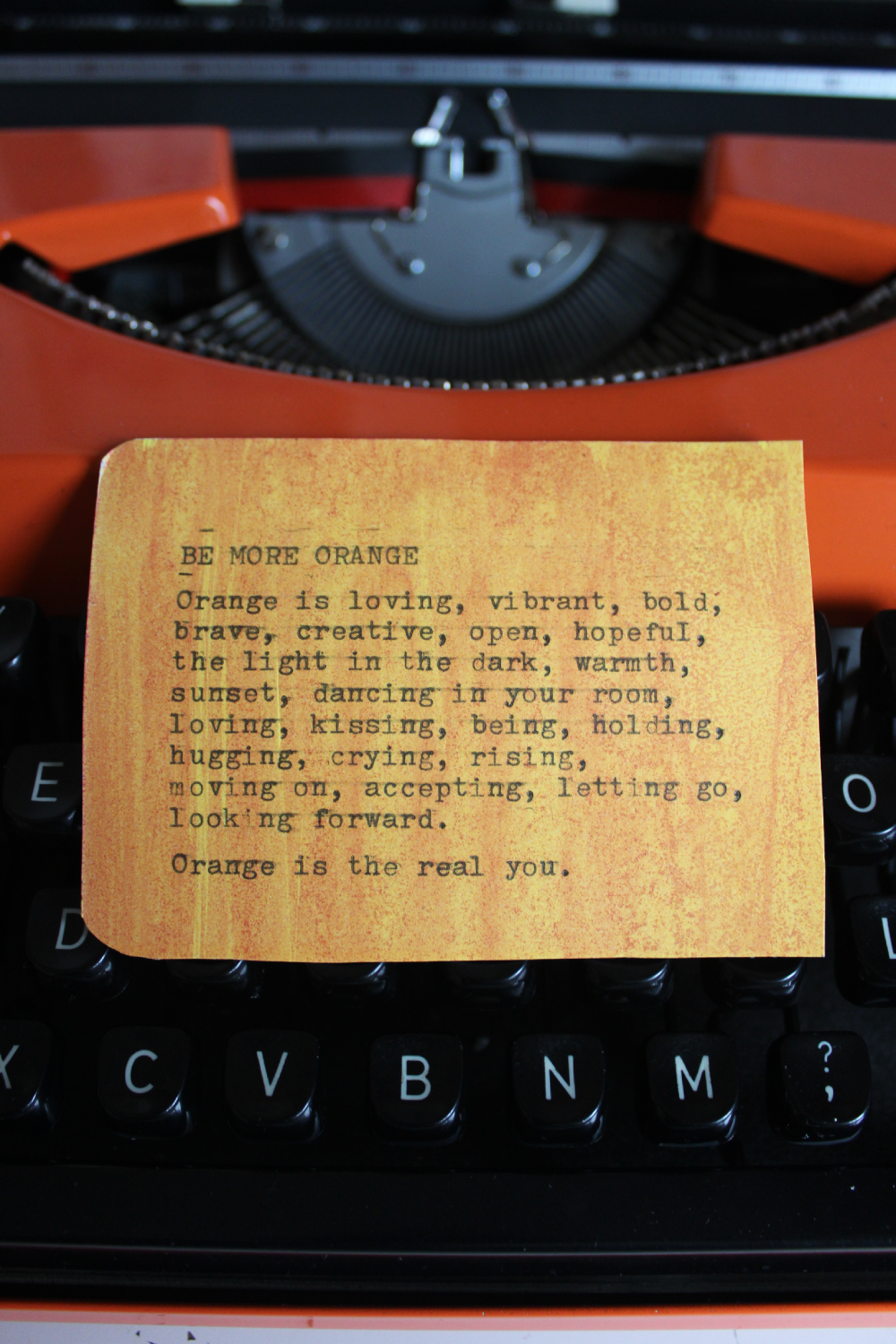 Close-up of a vintage orange typewriter with a yellow paper typed with the poem "Be More Orange" placed on the keys.