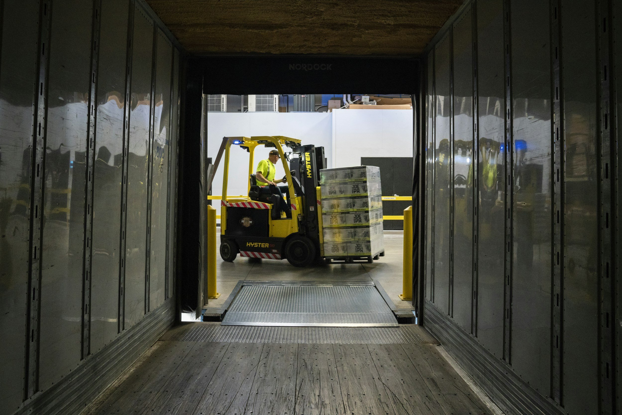 Looking through a tunnel at a warehouse loading dock, a worker on a yellow forklift is moving pallets of products outside.