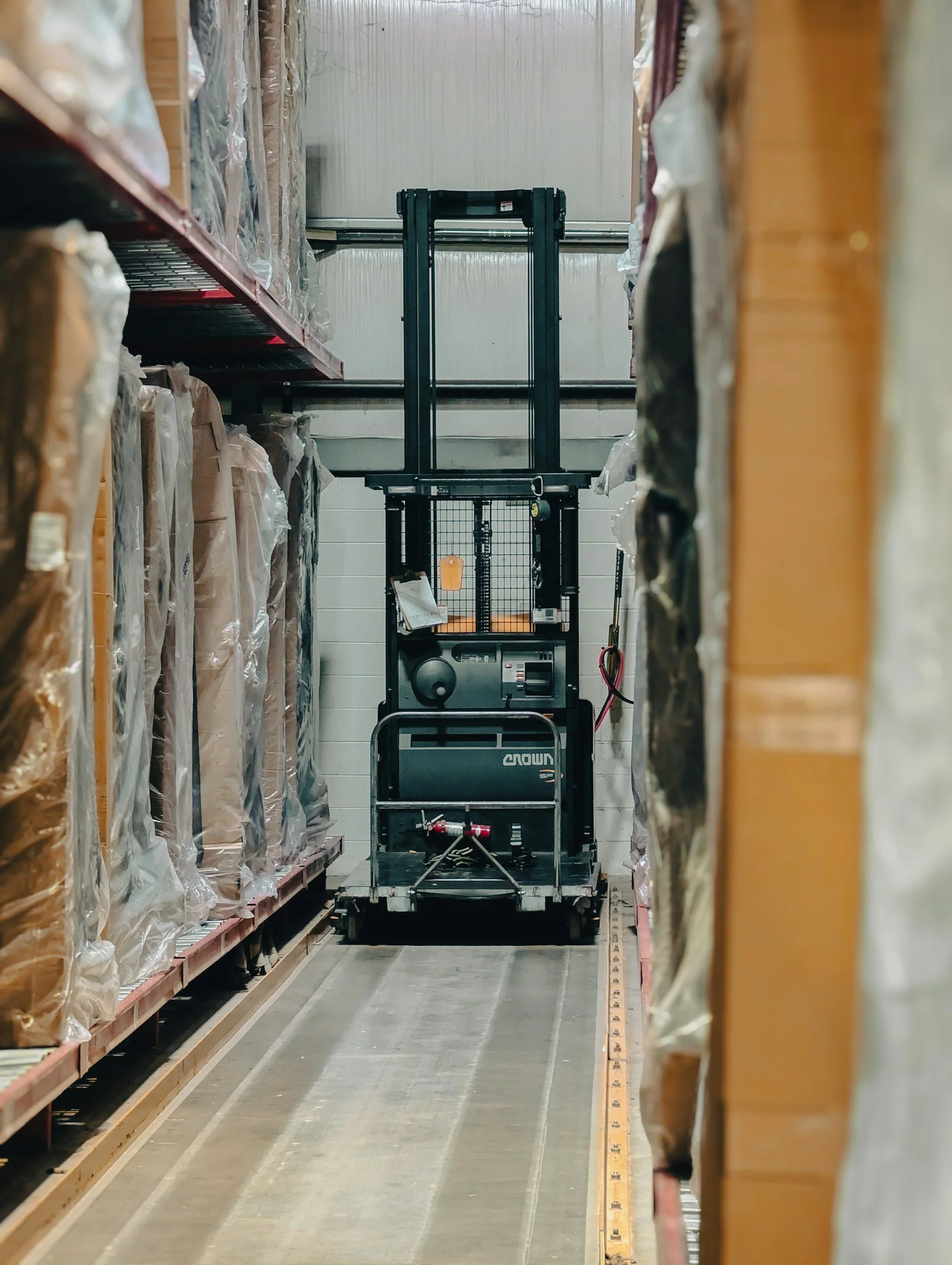 A narrow aisle in a warehouse with a black electric pallet jack in the center, surrounded by shelves stocked with items wrapped in plastic on both sides.