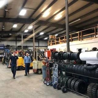 Warehouse interior with two workers walking among shelves of materials and equipment, including large rolls of black tubing and a yellow forklift.