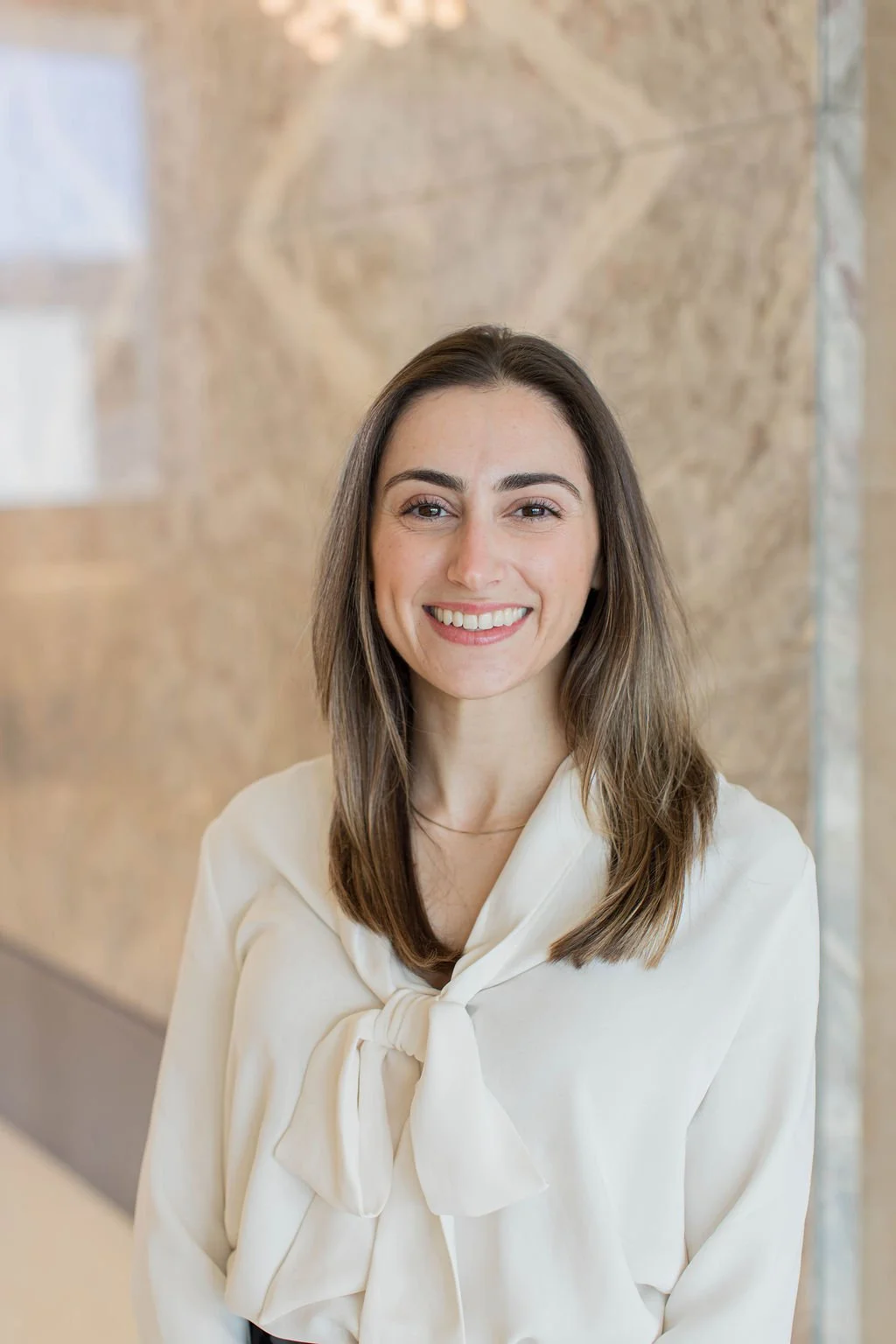 A woman with long brown hair smiling at the camera, wearing a white blouse with a large bow at the neck, standing indoors against a marble wall background.