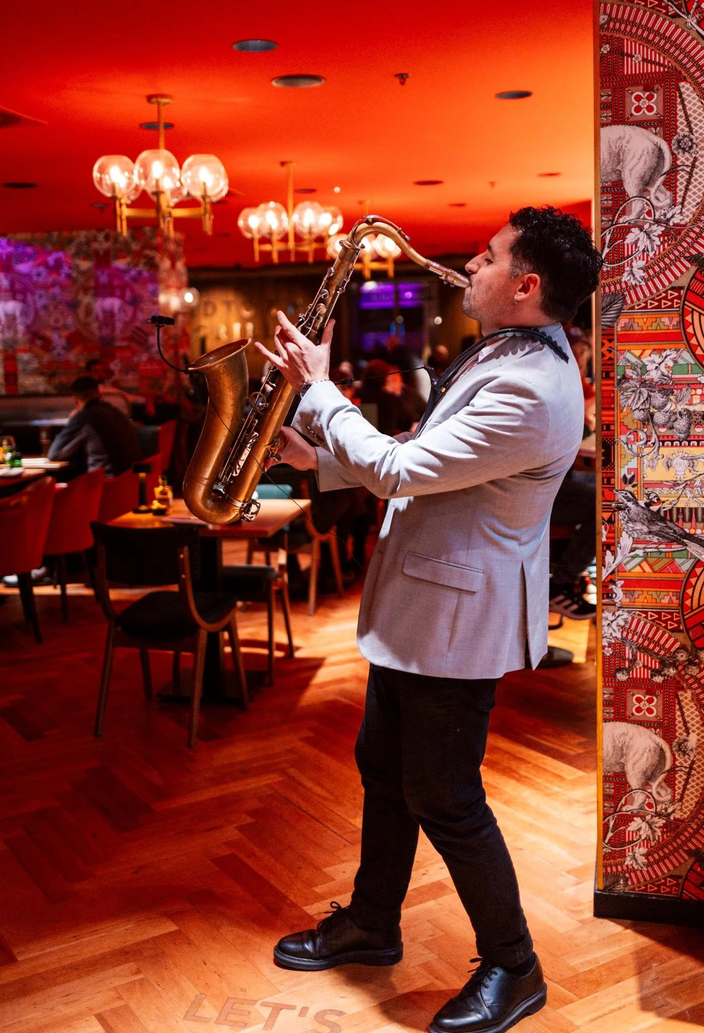 A man in a light gray blazer and black pants playing a saxophone in a dimly lit restaurant or bar with red ceiling lights, surrounded by tables and patrons.