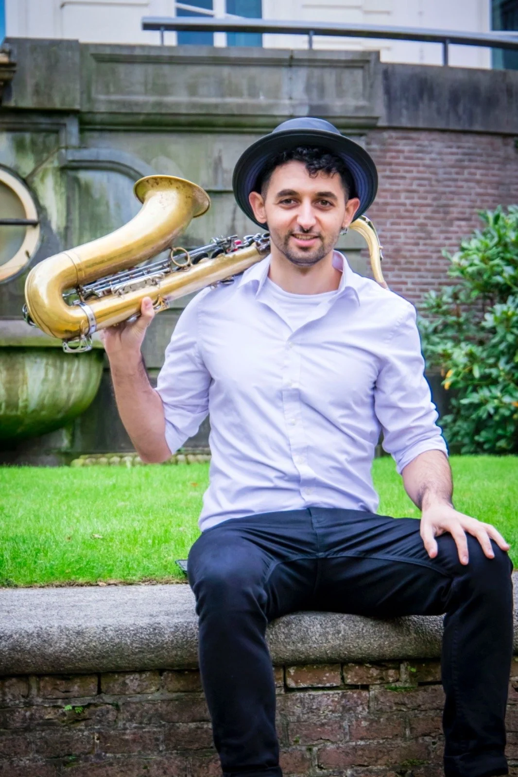 A young man with dark curly hair, wearing a black hat and a white shirt, sitting on a stone bench outdoors. He is smiling and holding a saxophone on his shoulder. The background includes a grassy area, a brick wall, and some green bushes.