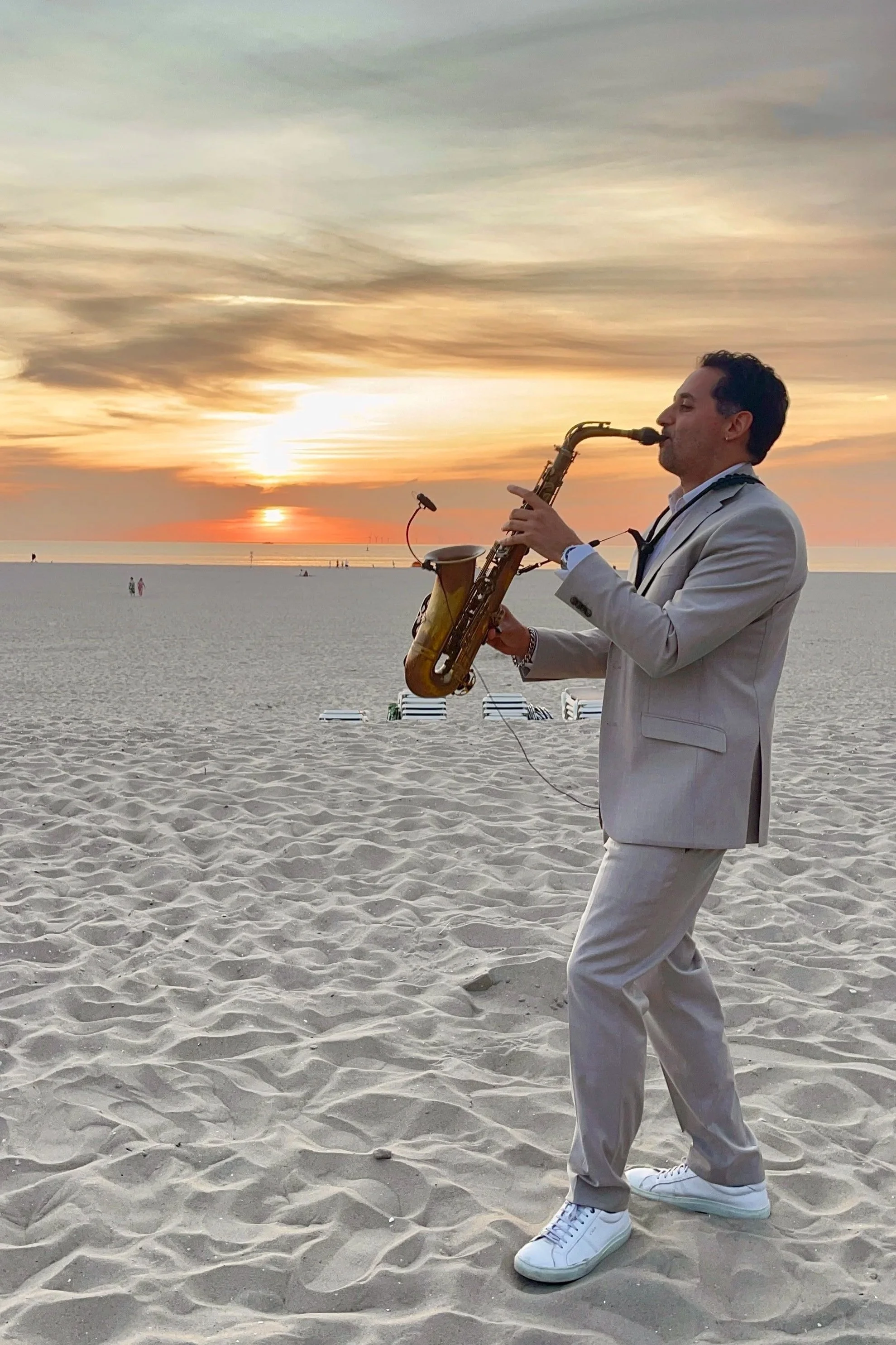 A man in a beige suit playing a saxophone on the beach during sunset.
