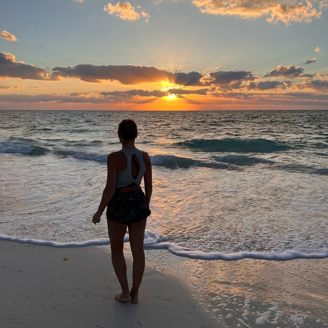 Eine Frau steht am Strand und schaut auf das Meer während eines Sonnenuntergangs mit bewölktem Himmel.
