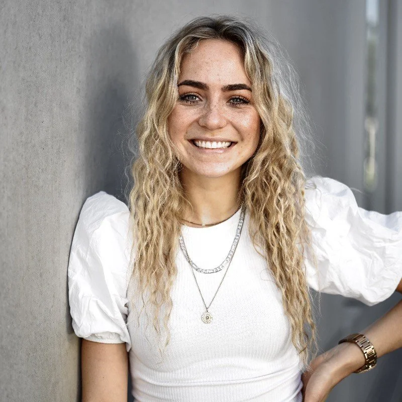 Lächelnde junge Frau mit blonden, lockigen Haaren, trägt weiße Bluse mit Puffärmeln, mehrere Silberketten, Armbanduhr, steht an einer Wand und lächelt.